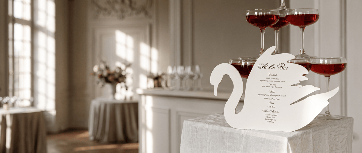 A white swan-shaped menu card reading 'At the Bar' displayed on a white tablecloth in a softly lit event venue, with champagne flutes and tables blurred in the background.
