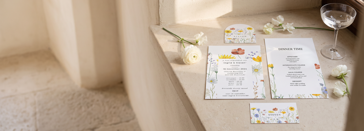 A styled flat-lay of a wedding invitation suite on a stone windowsill, featuring floral stationery cards, a program, a small card, and a wine glass.
