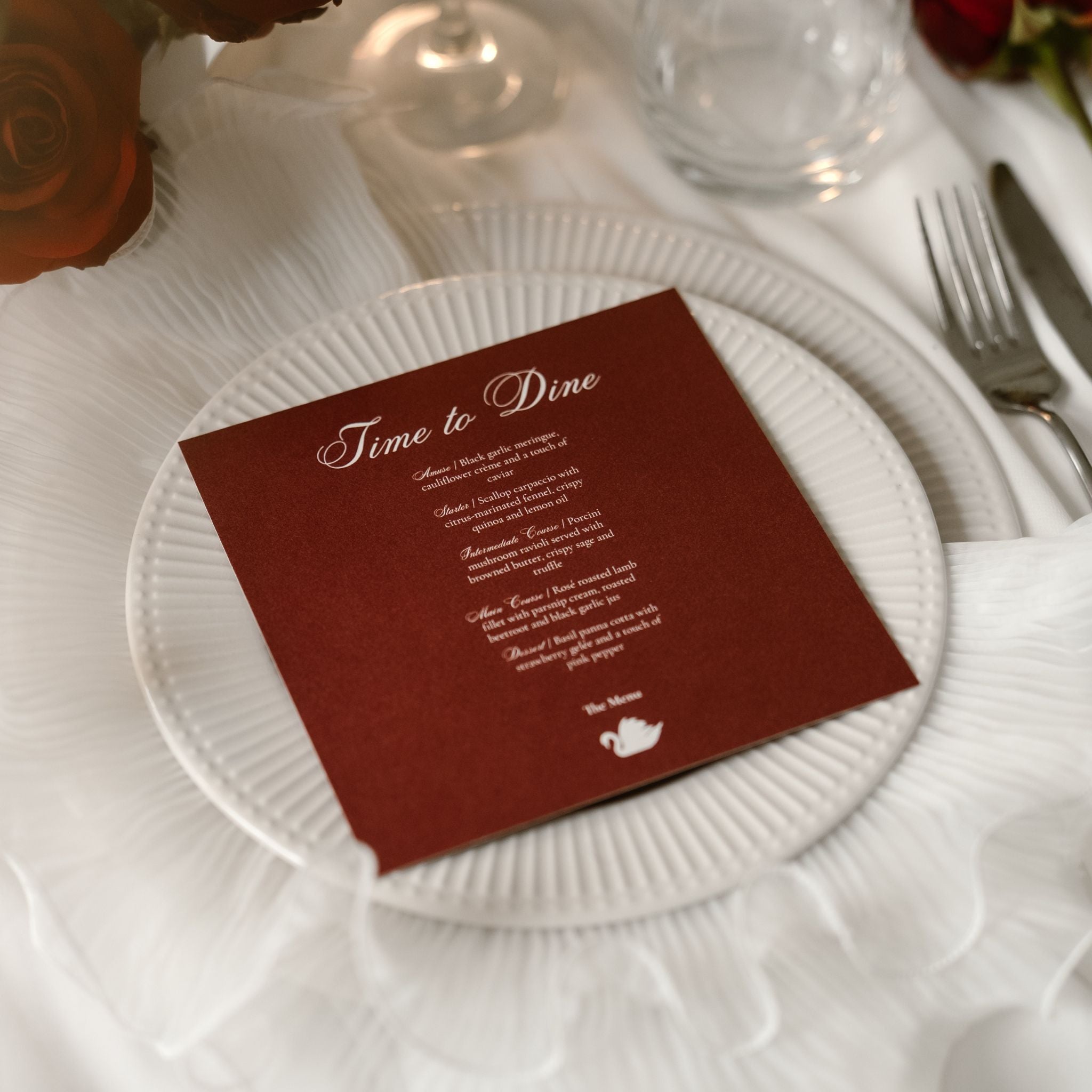A square burgundy menu reading 'Time to Dine' with white script and text, styled on a white plate over a white tablecloth with a fork, knife and glass nearby.