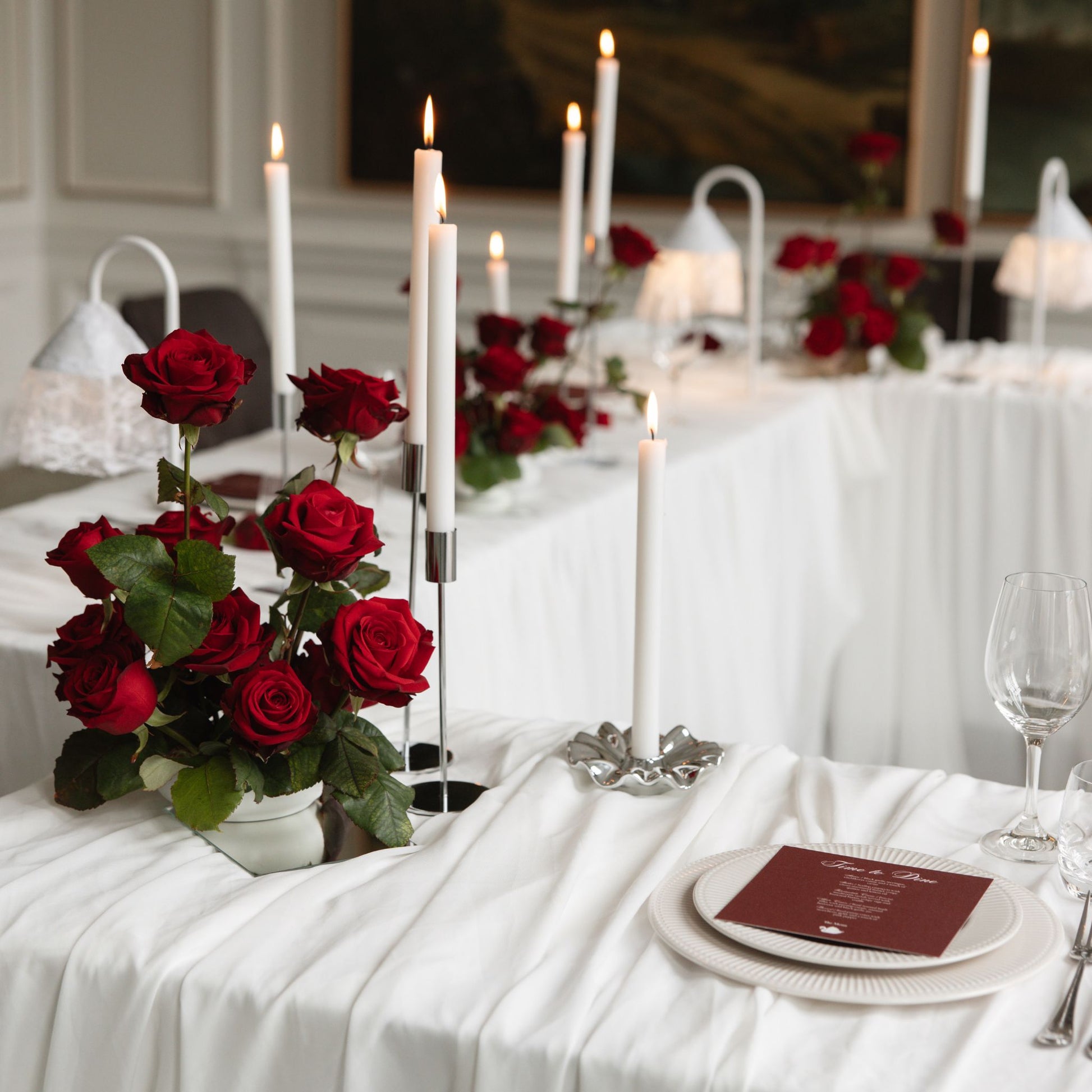 A formal event table setting featuring a vibrant red rose centerpiece in a white vase, tall white taper candles in silver holders, white table linens, and a bordeaux menu on a white plate.