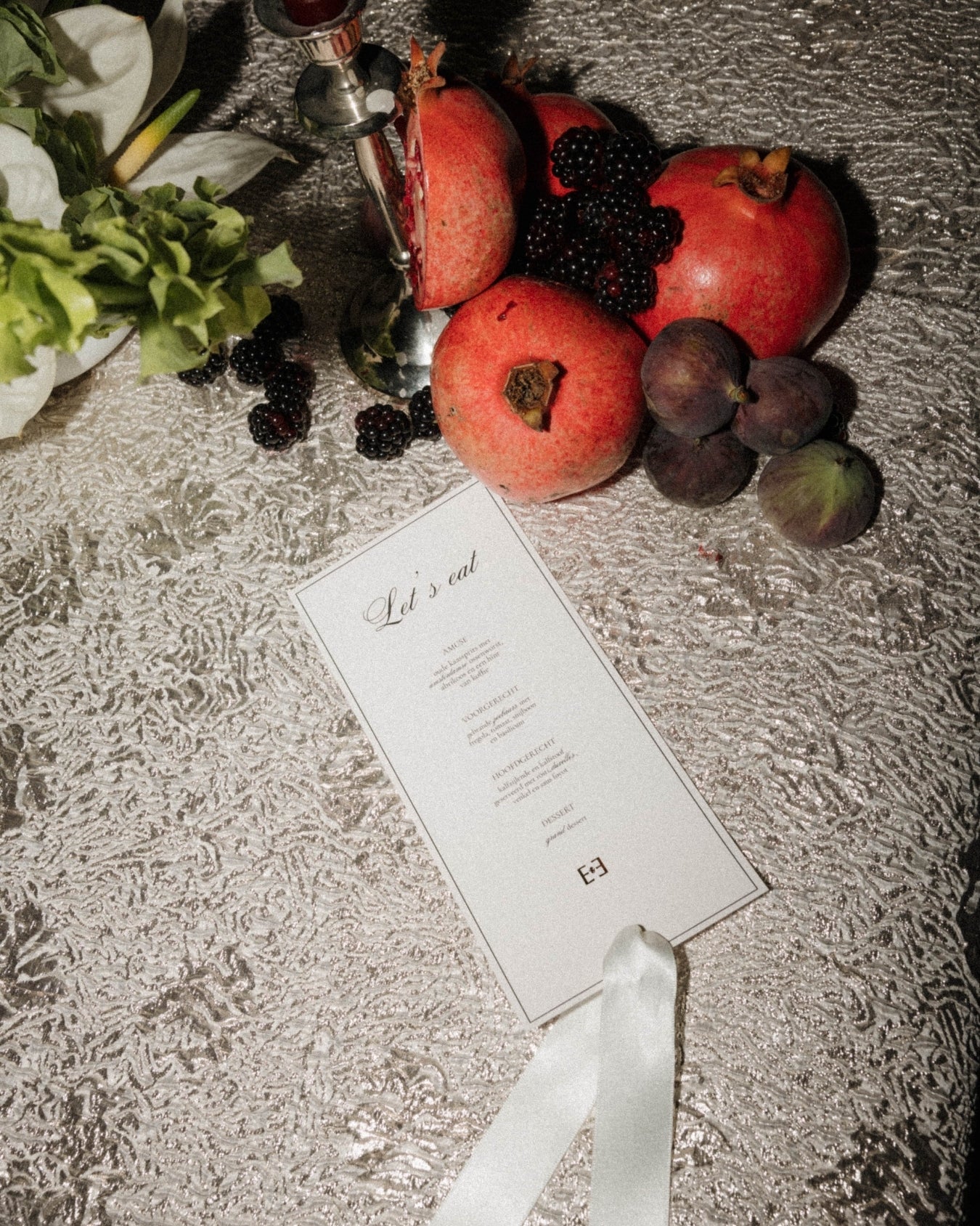 Fruit arrangement with pomegranates, blackberries, and a silver teapot on a textured surface with a 'Let's eat' card.