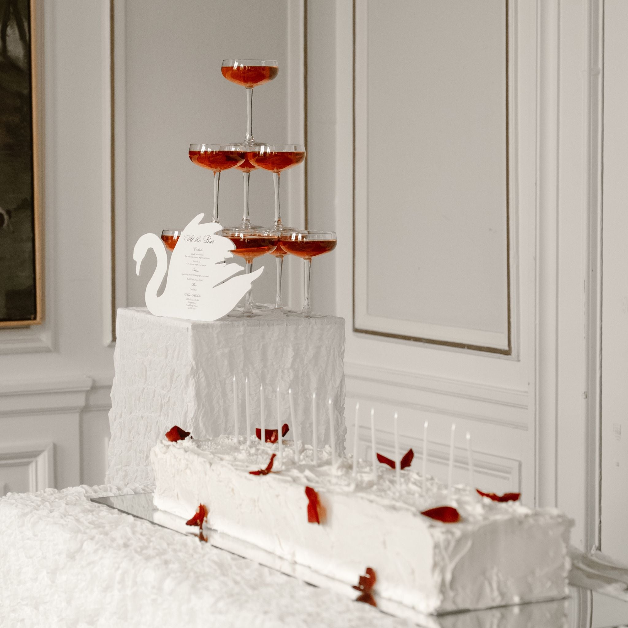 A white tiered wedding cake with candles and red rose petals, styled with a champagne tower of six stemmed glasses on a white pedestal in an elegant room.