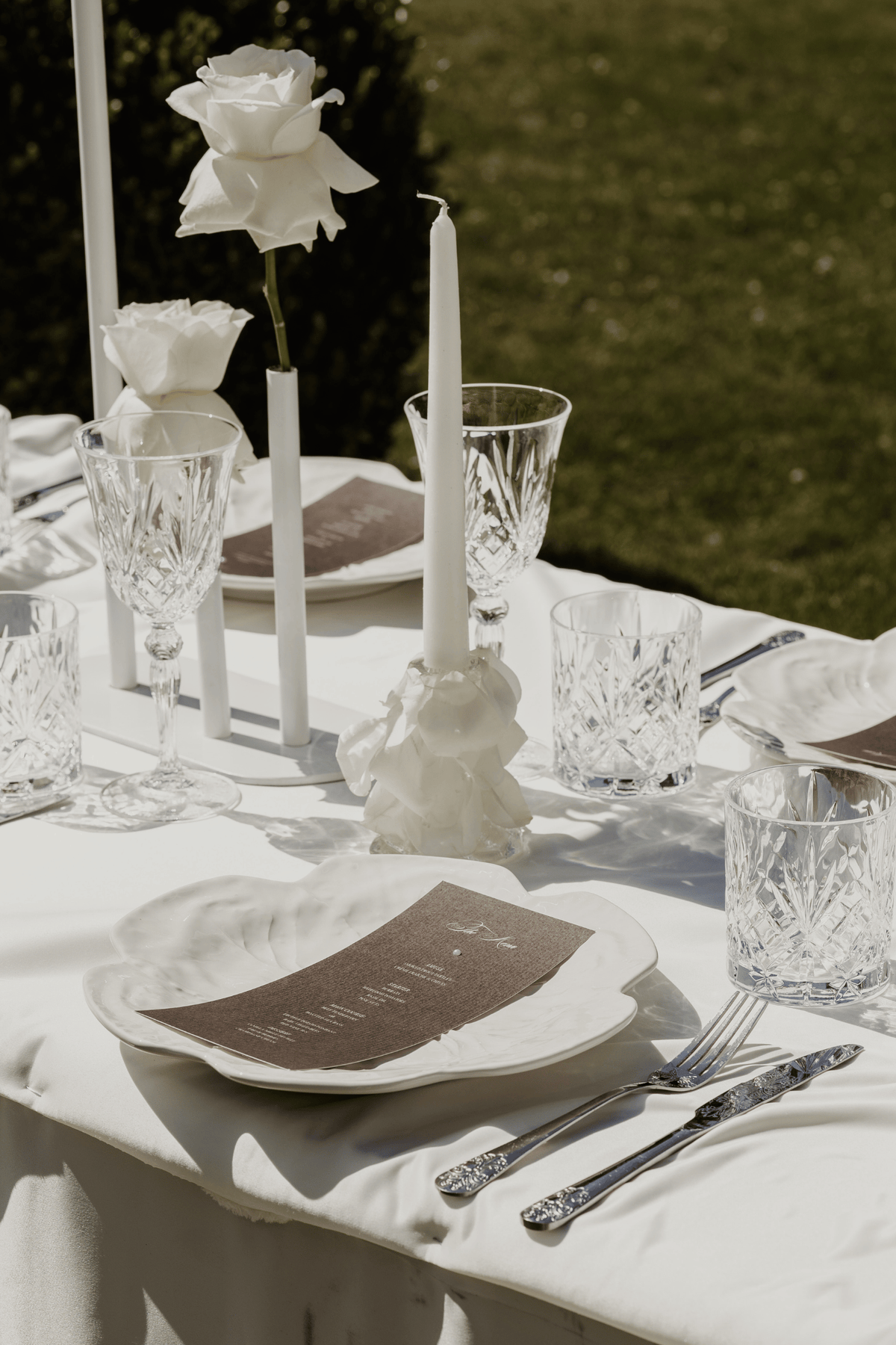 An elegant outdoor table setting with white tablecloths, white ceramic plates, silver flatware, clear cut-glass water glasses, white taper candles, and white roses, photographed in natural sunlight with a green lawn in the background.