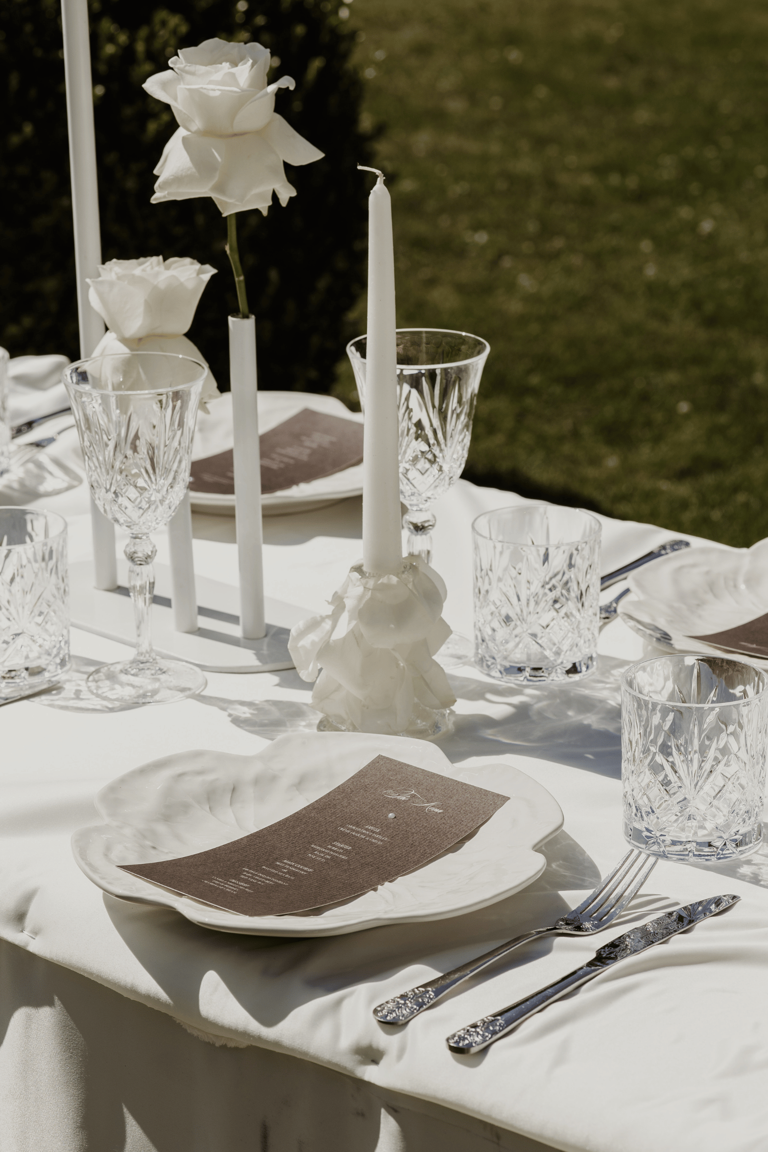 An elegant outdoor table setting with white tablecloths, white ceramic plates, silver flatware, clear cut-glass water glasses, white taper candles, and white roses, photographed in natural sunlight with a green lawn in the background.