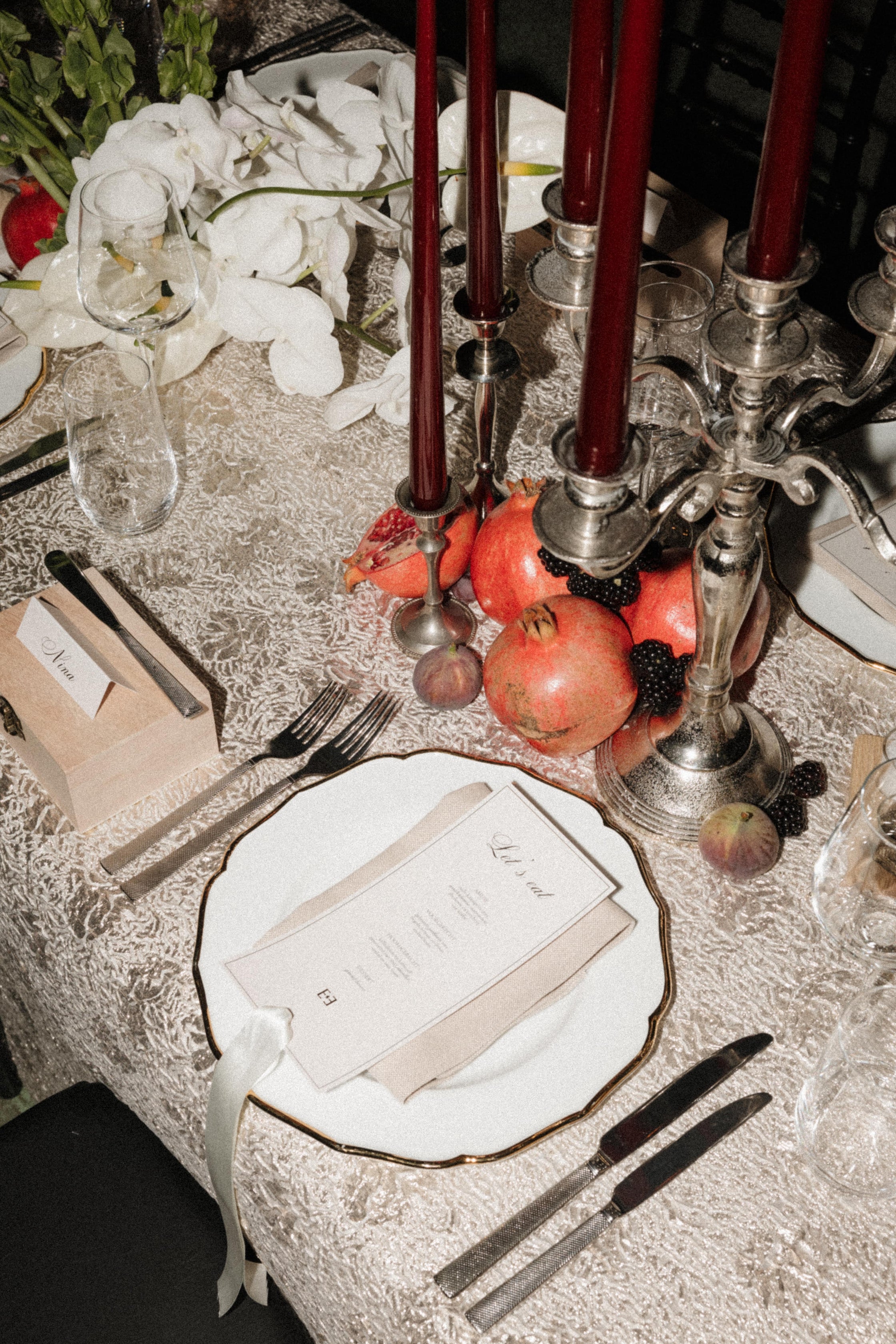 A styled table setting with a white plate, silver cutlery, a beige napkin, dark red taper candles in silver candelabras, pomegranates, blackberries, and white flowers on a textured tablecloth.