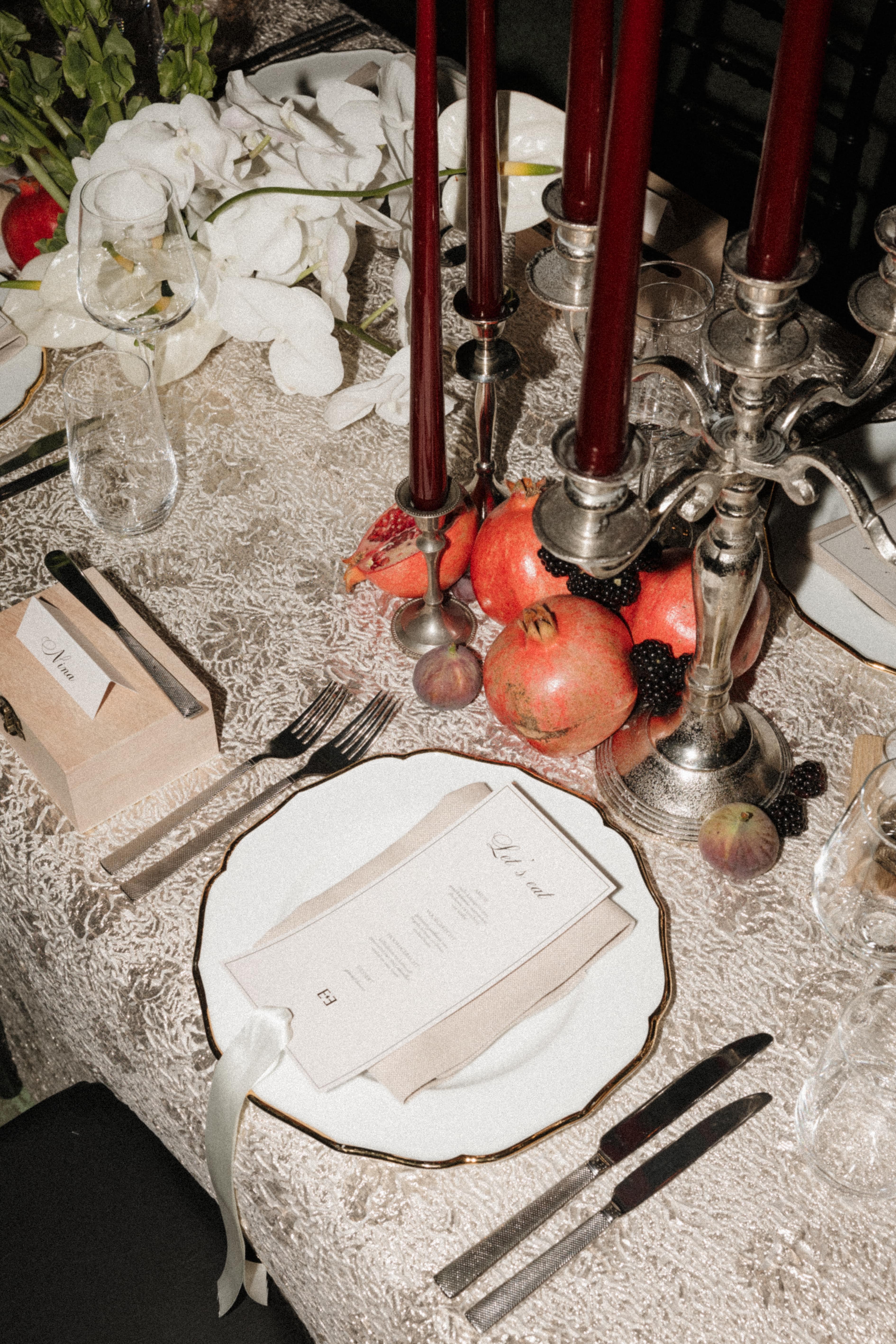 A styled table setting with a white plate, silver cutlery, a beige napkin, dark red taper candles in silver candelabras, pomegranates, blackberries, and white flowers on a textured tablecloth.