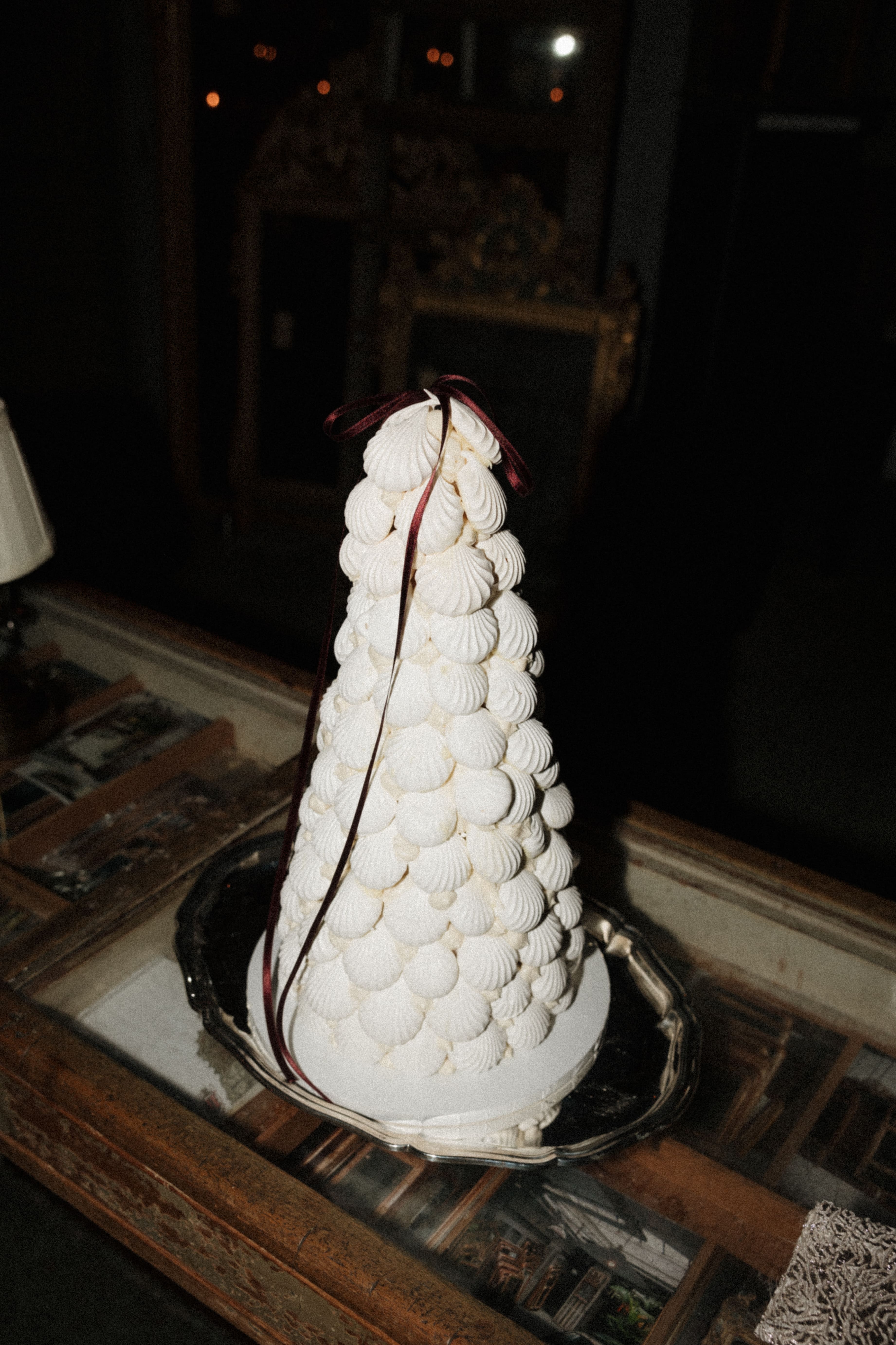 A white, cone-shaped decorative cake or pastry covered in piped meringue-like dollops, topped with a dark red ribbon, displayed on a silver tray on a wooden table in a dimly lit room.