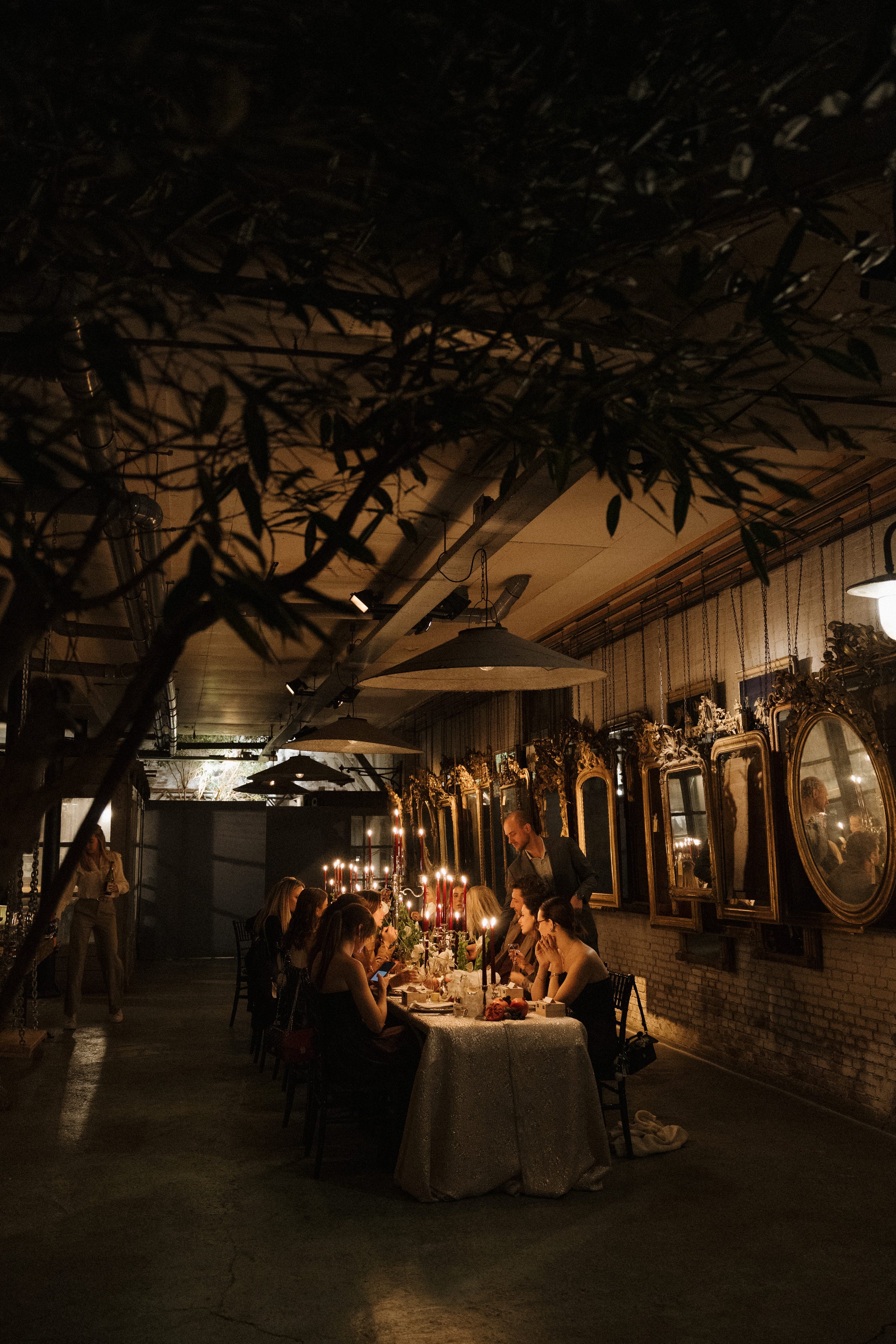 A dimly lit dining scene in a rustic indoor venue, with a group of people seated around a table covered in a white tablecloth and lit by candles. Large dark tree branches hang overhead, and the wall behind features mirrors and warm ambient lighting.