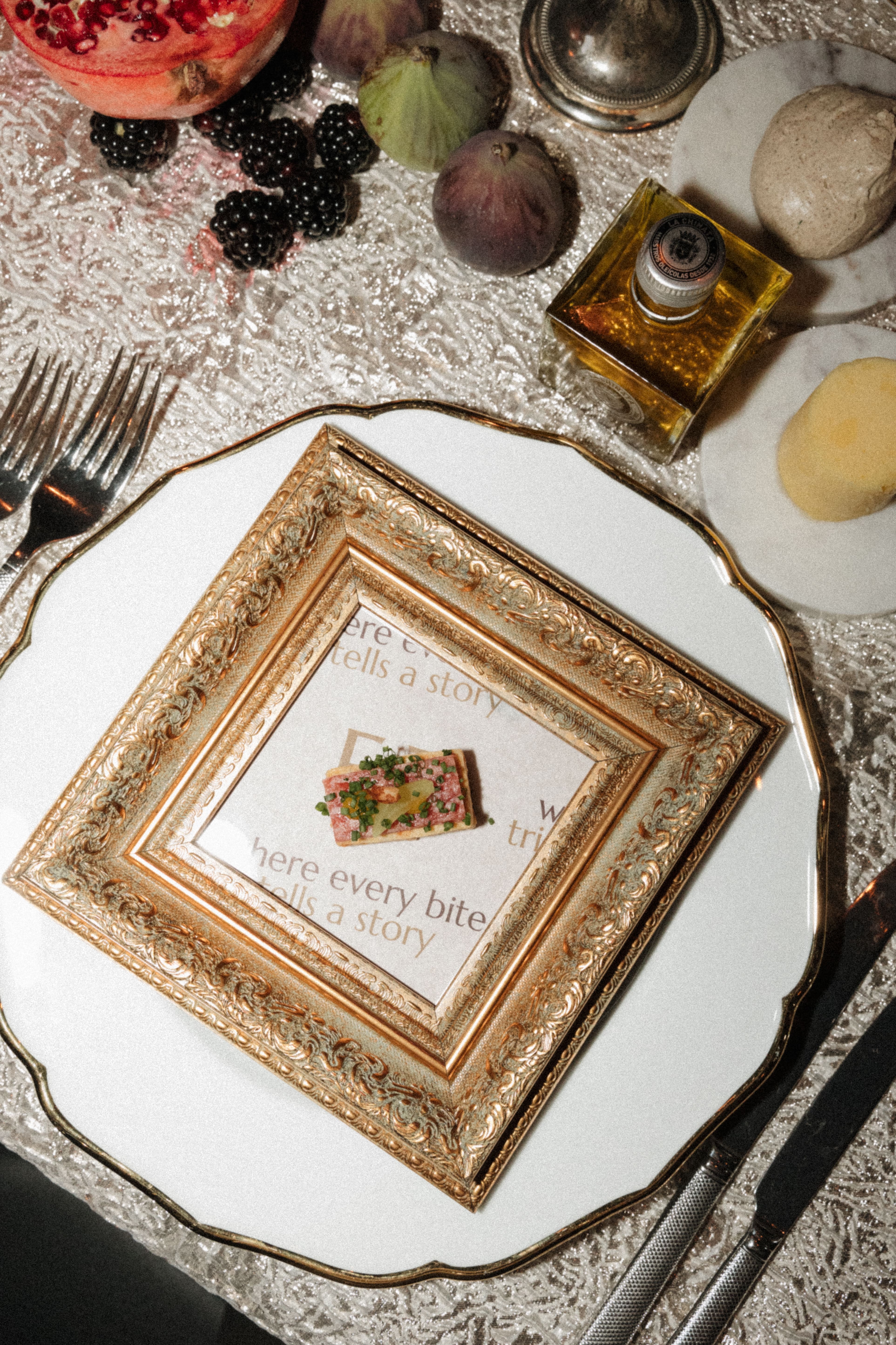 An ornate gold and white decorative frame holding a printed card with a small illustrated sandwich, arranged on a white plate at a styled table setting with cutlery, fruit, and a small bottle of oil.