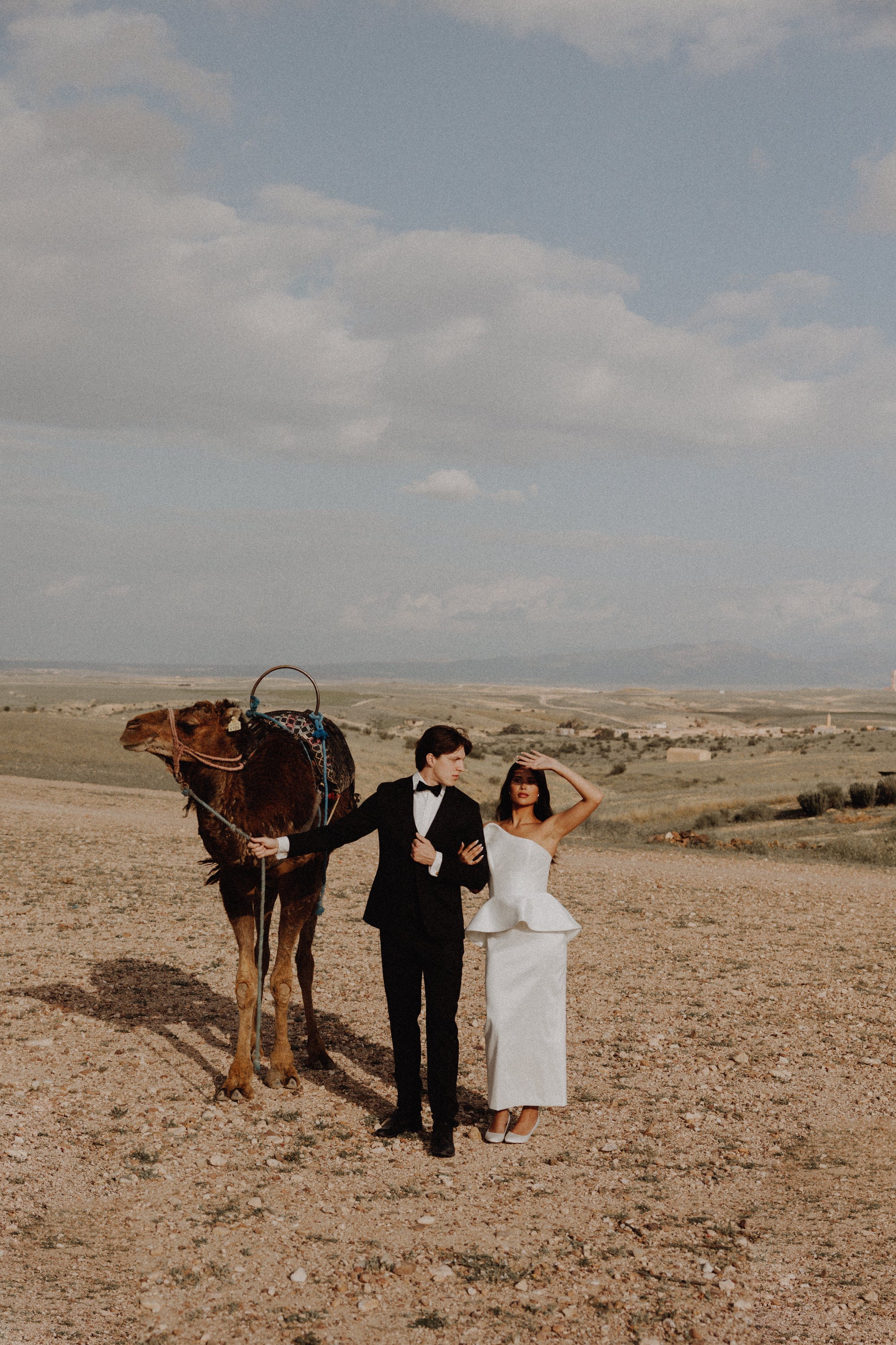A bride in a white sleeveless peplum wedding gown and a groom in a black tuxedo stand beside a camel in a desert landscape under a cloudy sky.