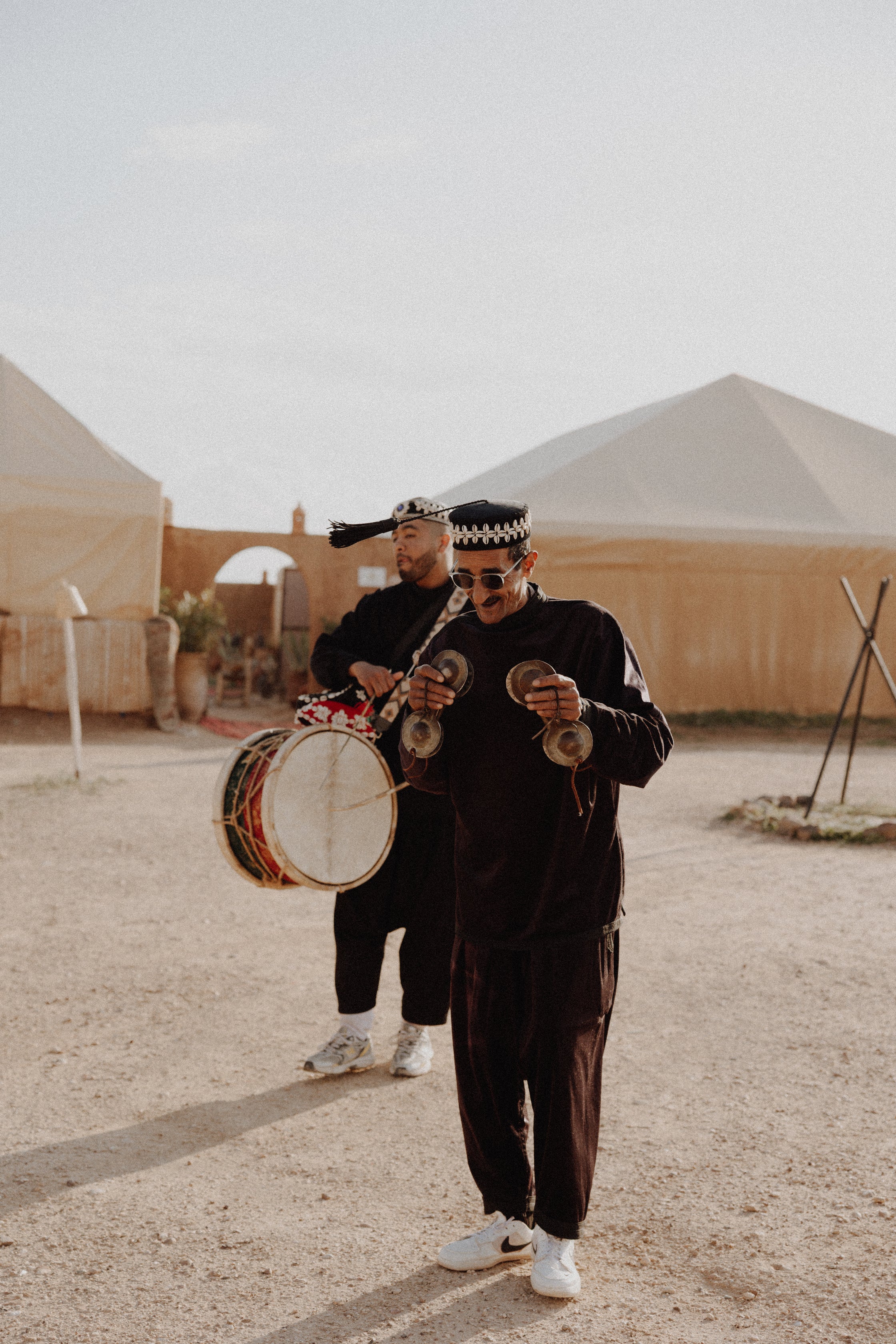 Two men in black traditional attire playing musical instruments outdoors in a desert setting with tents in the background.