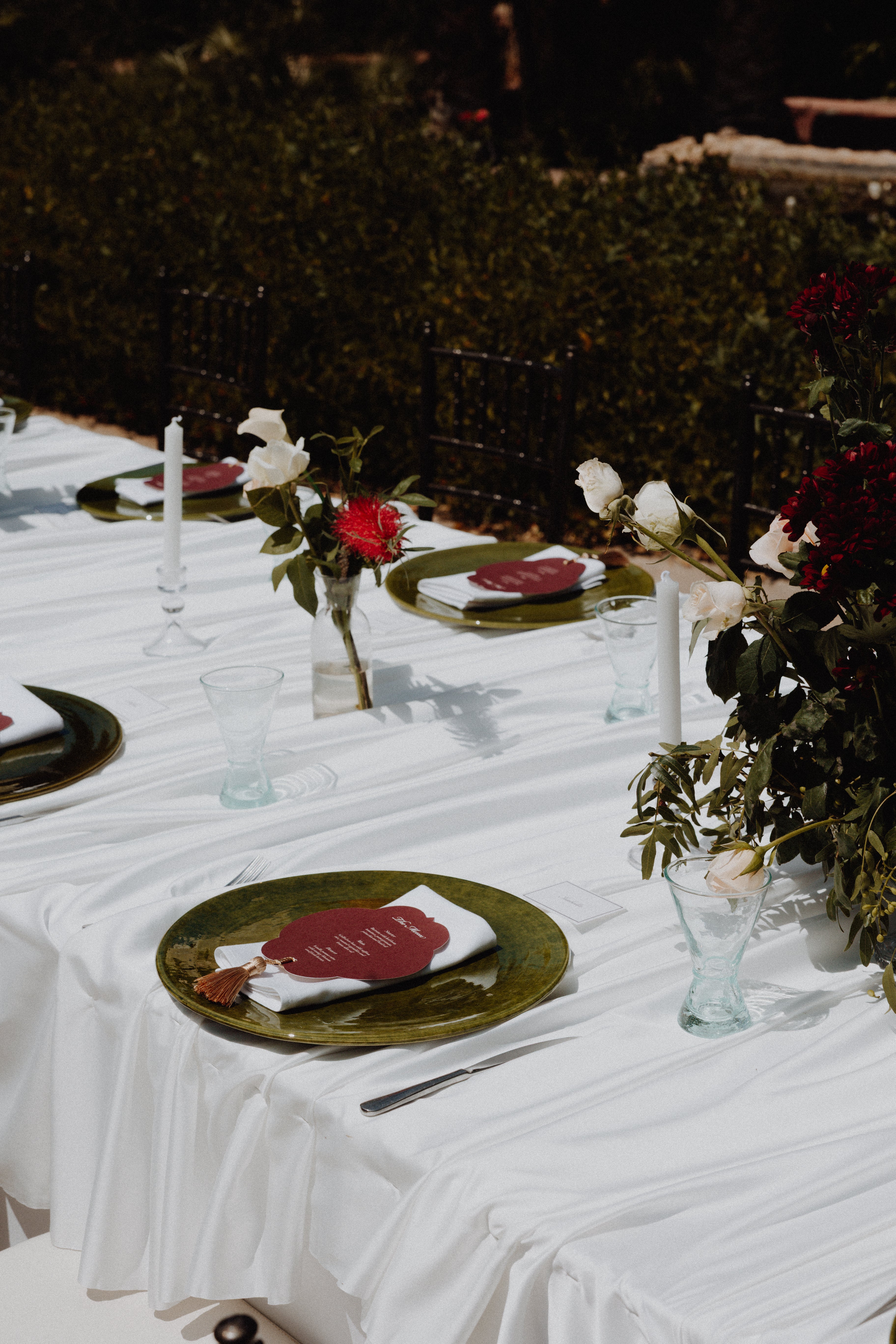 A styled outdoor wedding table setting with a white draped tablecloth, olive green plates, white and red roses, and clear glassware.
