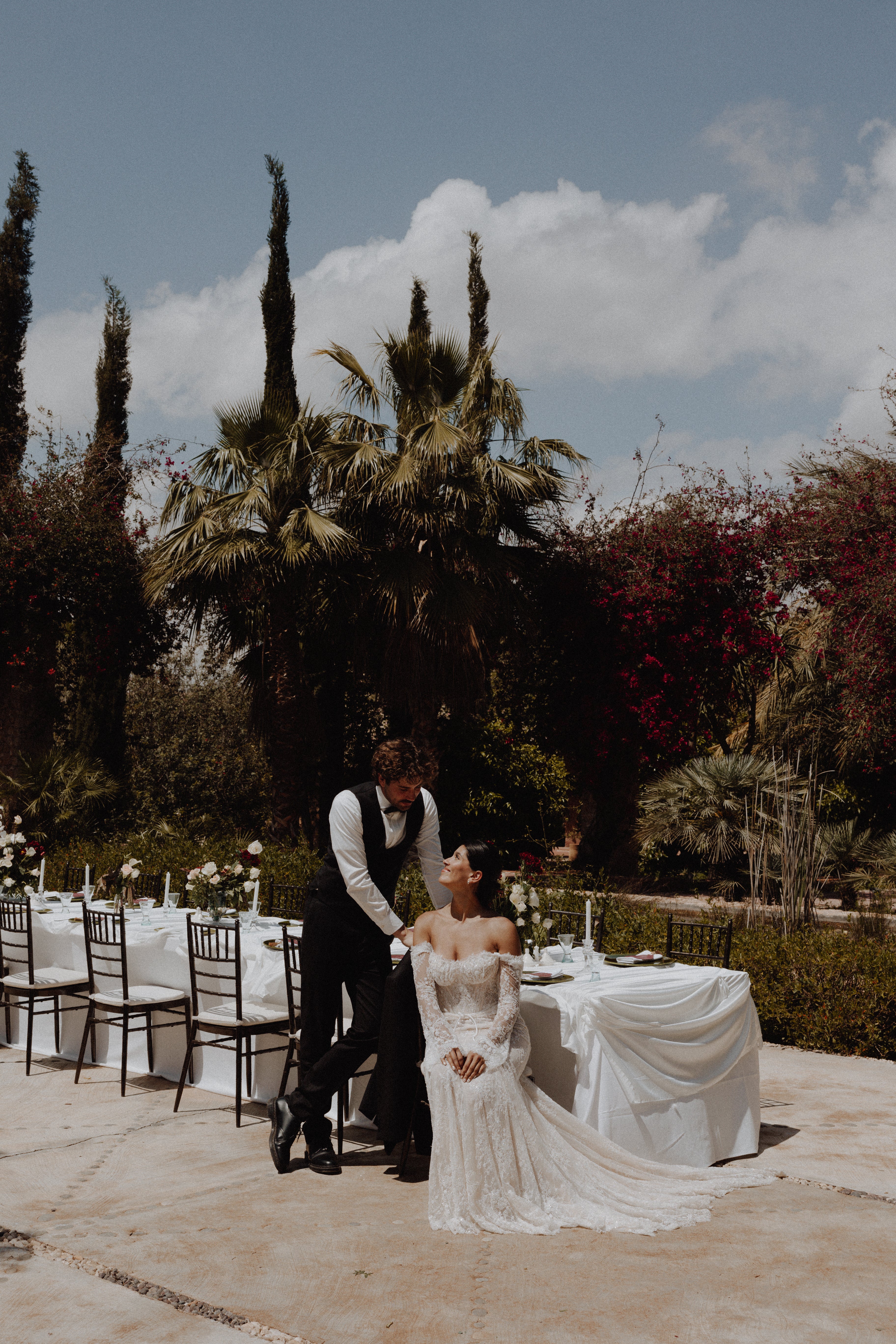A bride in an off-the-shoulder white wedding gown and a groom in a black vest and bow tie pose outdoors at a wedding reception, with tables, chairs, and palm trees in the background under a blue sky.