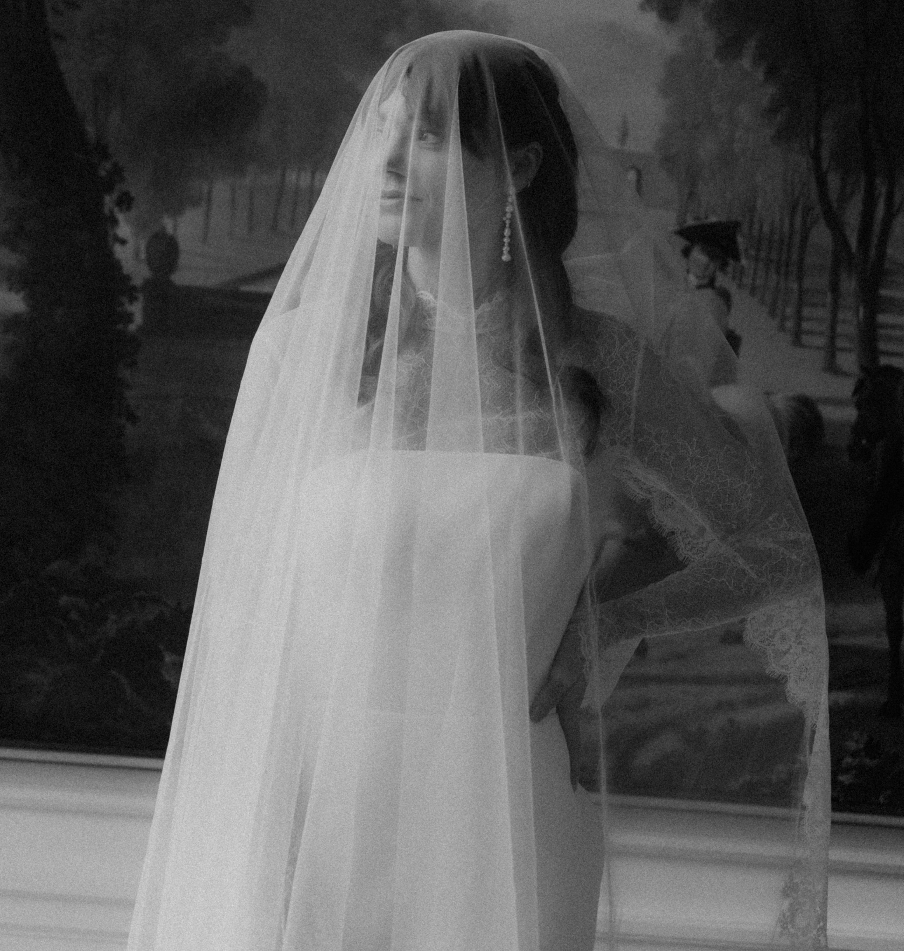 A woman wearing a long sheer bridal veil and a white wedding dress stands indoors in front of a large framed landscape painting. The photo is monochrome, and another figure appears faintly within the painting in the background.