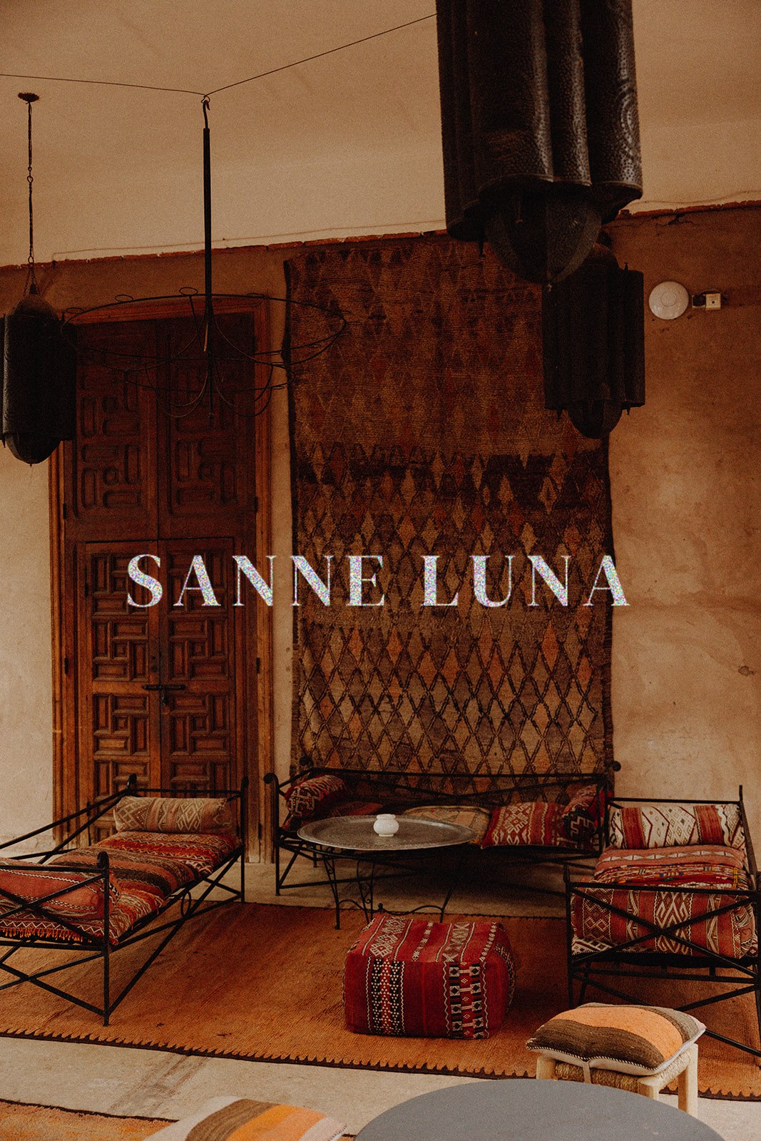 A styled interior scene with bohemian furniture and textiles, featuring a large patterned tapestry on the wall, two black metal lounge chairs with red patterned cushions, a small round coffee table, and a red patterned ottoman. The text 'SANNE LUNA' is overlaid across the center.