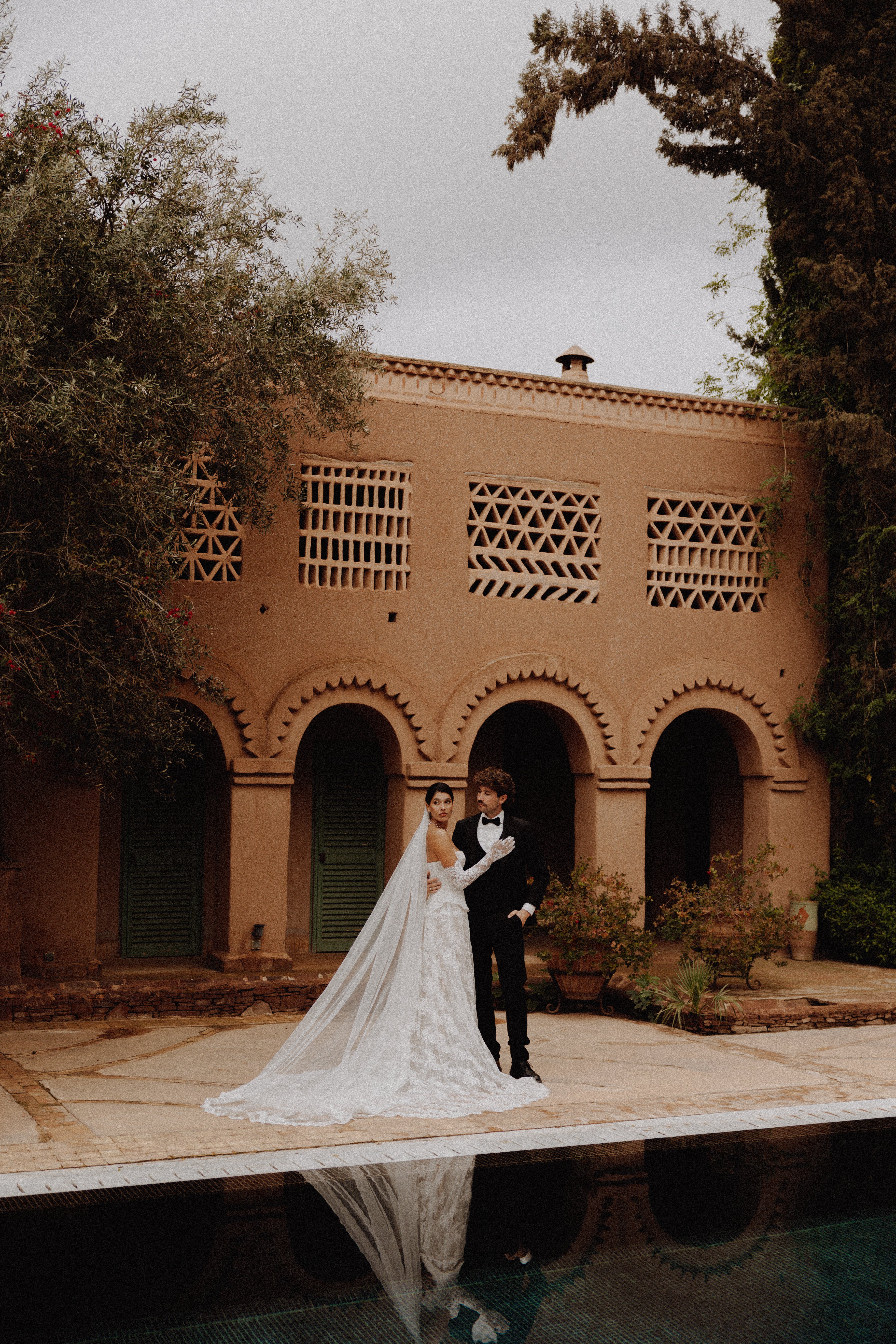A bride in a long white lace wedding gown with a sheer veil and a groom in a black suit pose in front of a traditional terracotta-colored courtyard building with arches, with a pool reflection below.