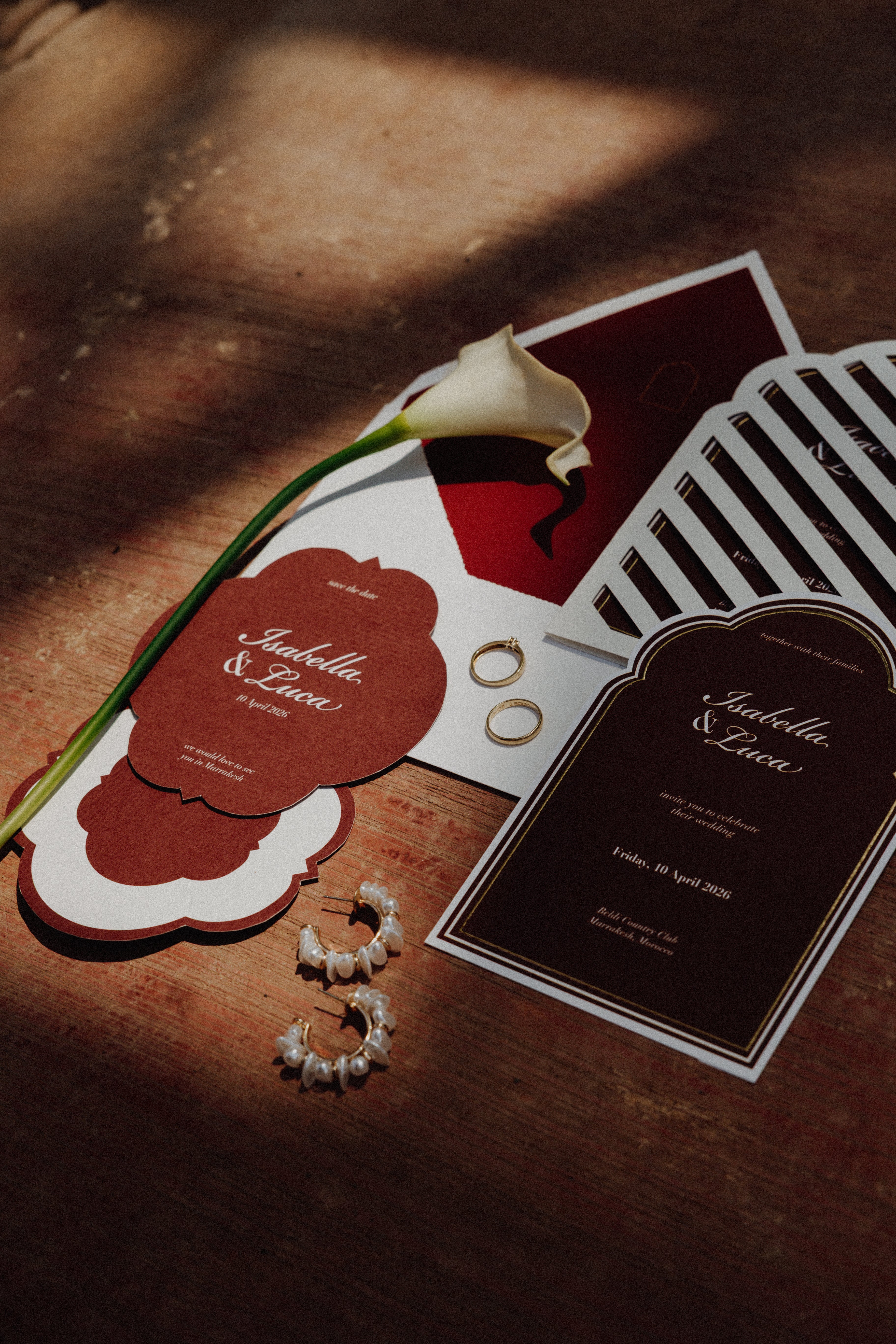 A styled flat-lay of wedding stationery on a wooden surface, including a dark red and white invitation card, a striped card, a calla lily flower, rings, and earrings. The lighting is dramatic with strong shadows.
