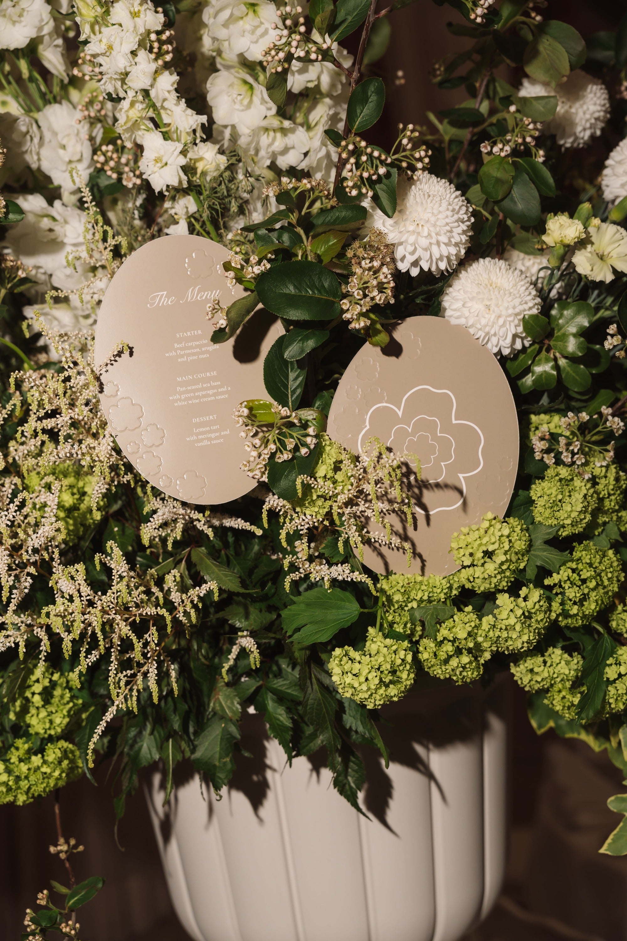 A close-up of a floral arrangement in a white ribbed vase, featuring white and green flowers with two round beige paper cards tucked among the leaves.