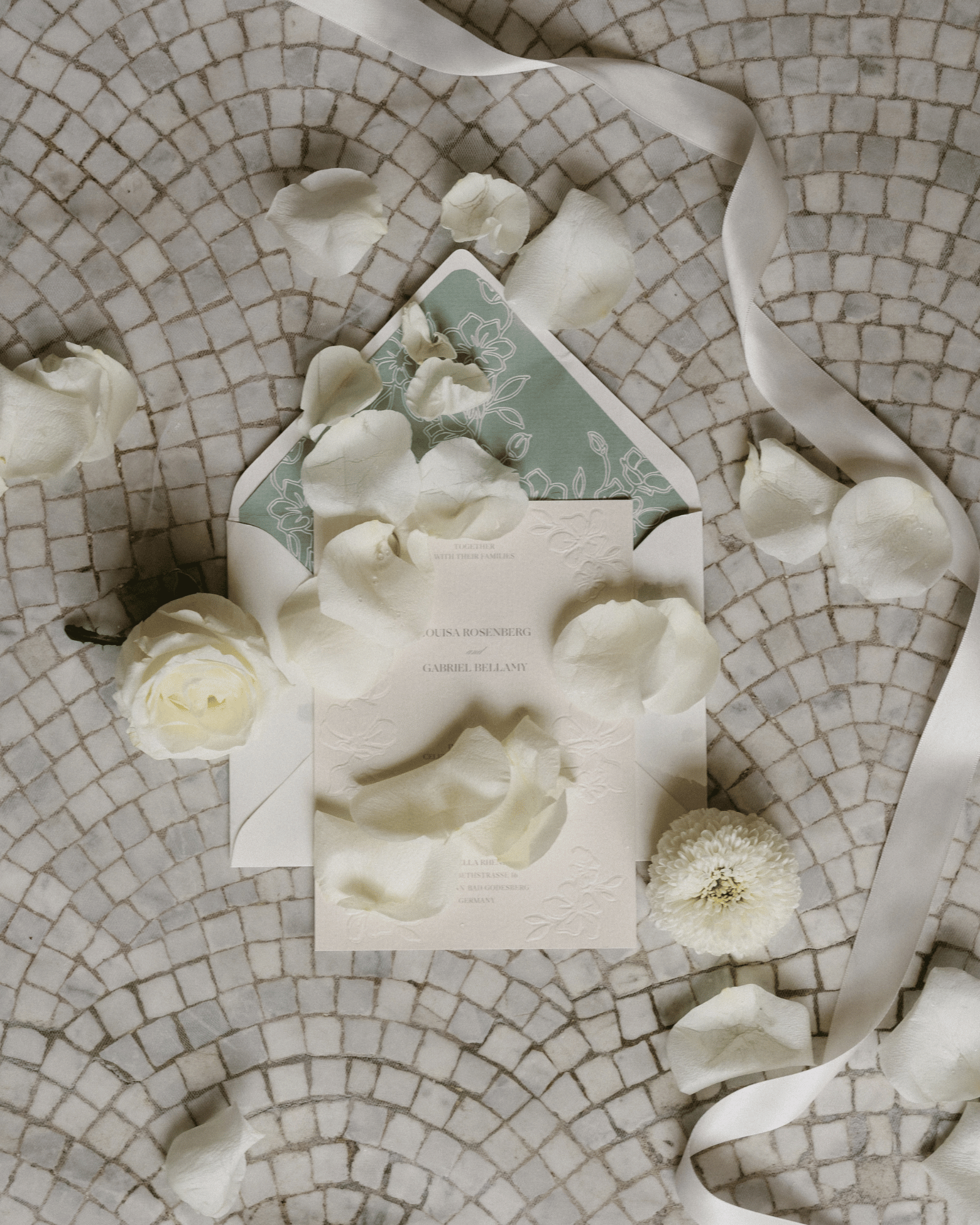 A styled flat-lay of a wedding invitation suite featuring a white card with embossed floral detailing, a sage green patterned envelope, and scattered white rose petals on a circular mosaic tile background with a white ribbon.