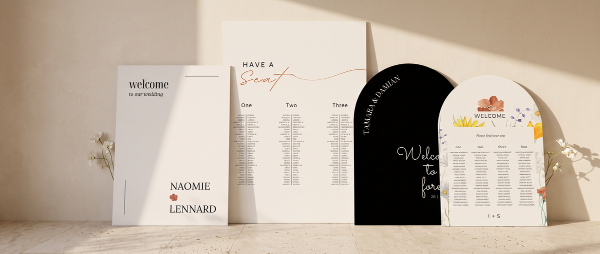 A coordinated stationery signage displayed on a tabletop against a warm beige wall. The set includes a white 'Welcome' card, a white seating chart, a black arch-shaped sign reading 'Welcome to our family,' and a white guest list with colorful floral accents.