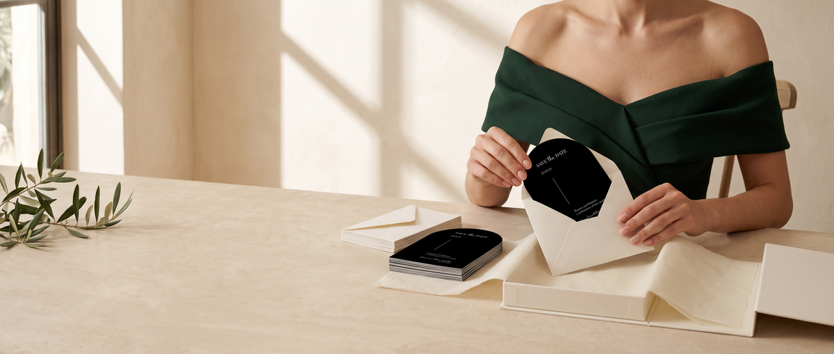 A woman in a dark green off-the-shoulder top sits at a light beige table, holding an open cream-colored envelope that contains a black card. A matching stack of black cards and cream envelopes lies on the table beside her, with soft window shadows and a small plant sprig in the background.