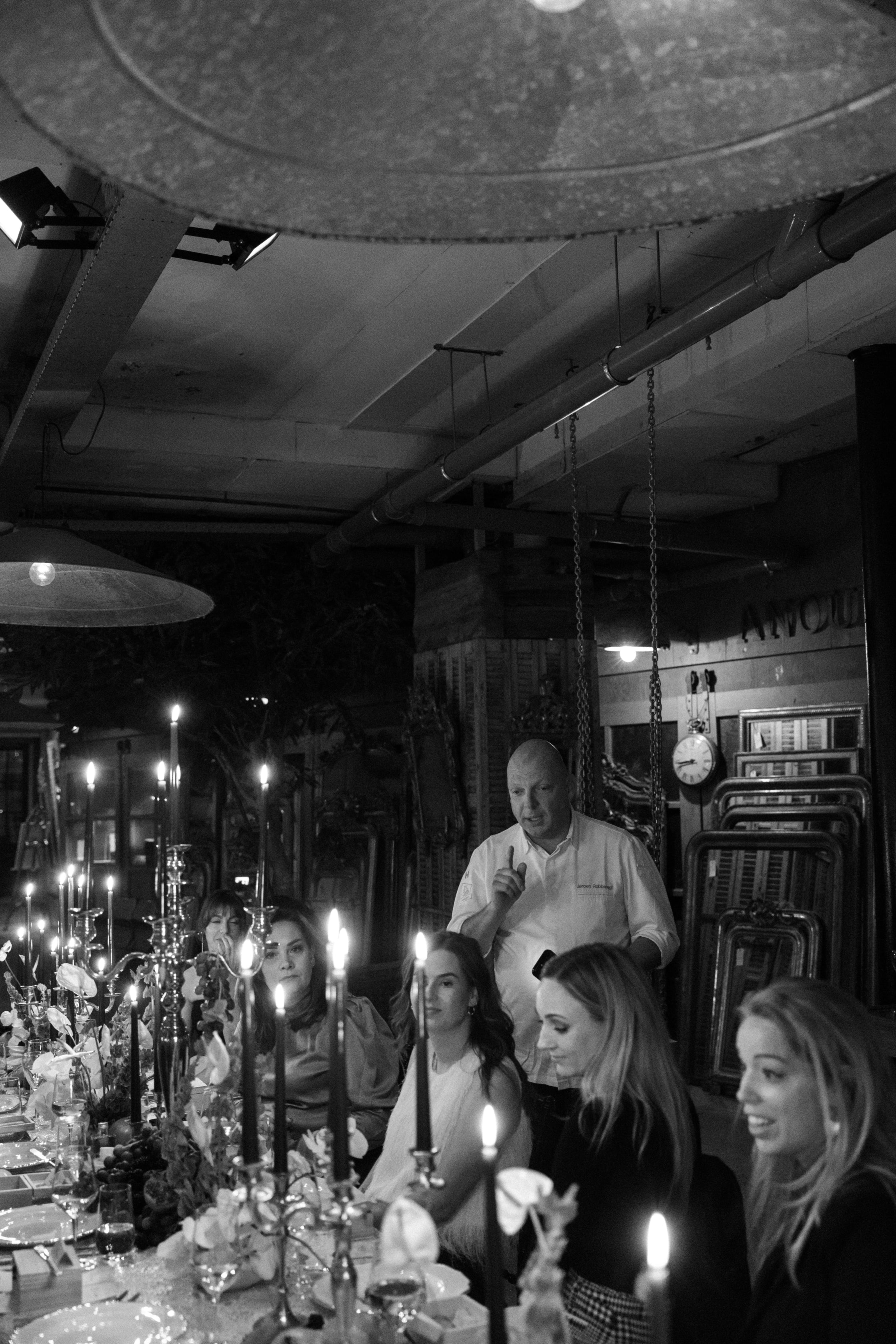Black and white photograph of a dimly lit dinner table scene with several people seated and standing around tall candlesticks and plates, under hanging industrial ceiling fixtures.