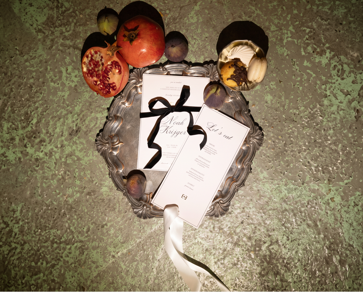 A styled flat-lay of printed invitation cards and a program on an ornate silver tray, surrounded by decorative fruits and a white ribbon, set against a textured green surface.