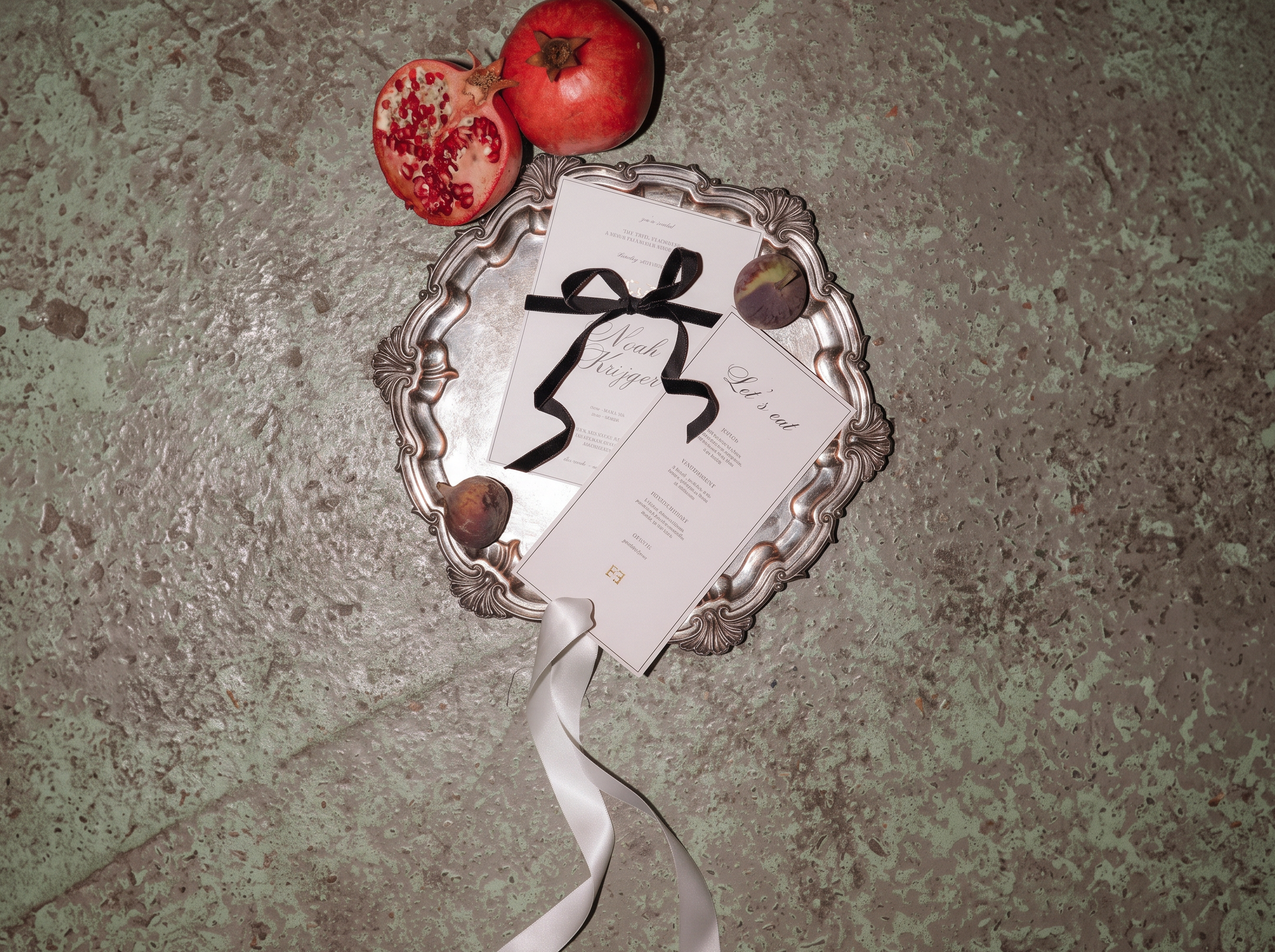 A styled flat-lay featuring a silver ornate tray holding a white invitation card with a black abstract signature, a white ribbon, and small acorns, accompanied by a whole and halved pomegranate on a textured green-gray stone surface.