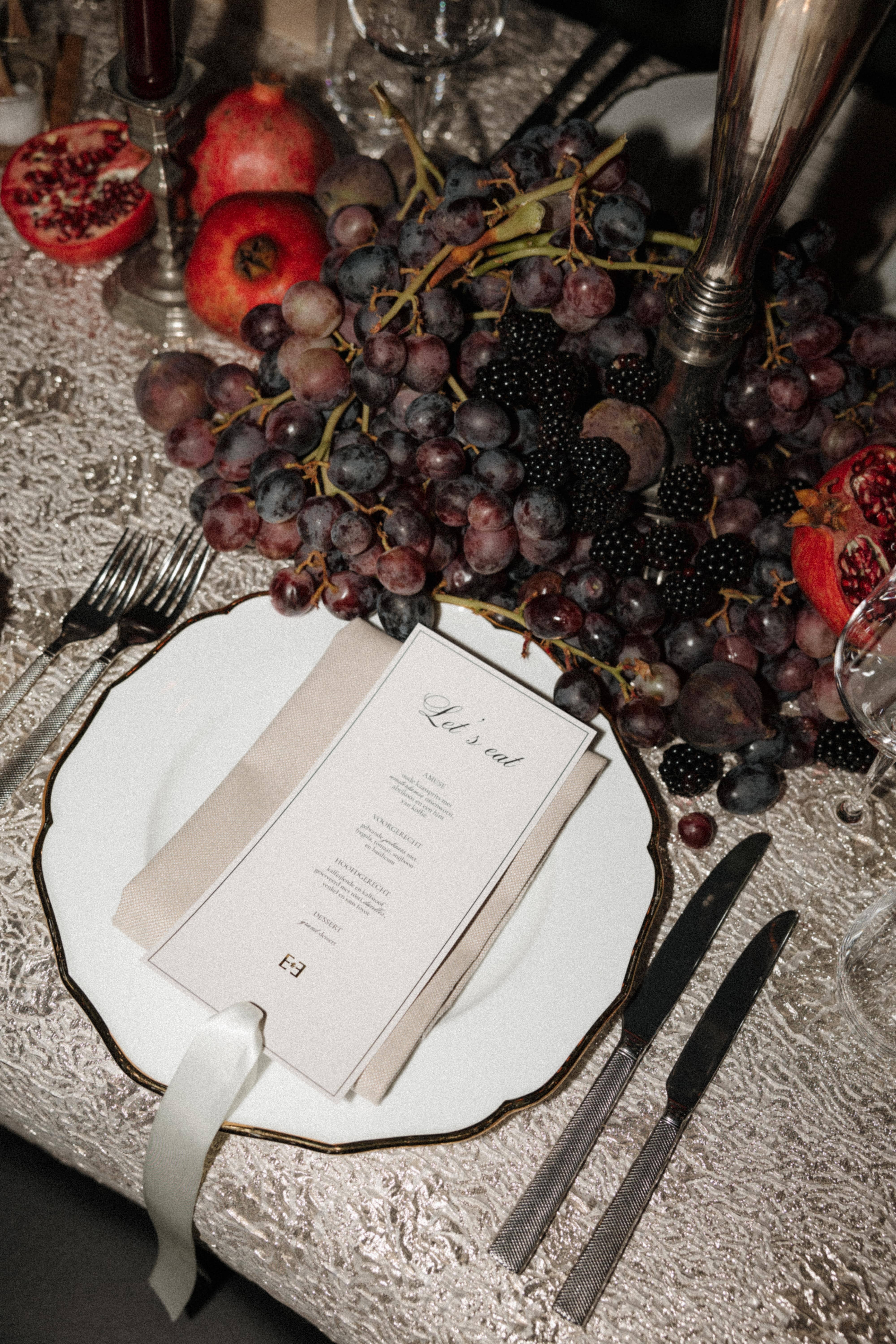 A styled table setting featuring a white scalloped plate with a gold rim, a beige napkin, a printed menu card, silver flatware, and a centerpiece of dark grapes, blackberries, and pomegranates on a textured white tablecloth.