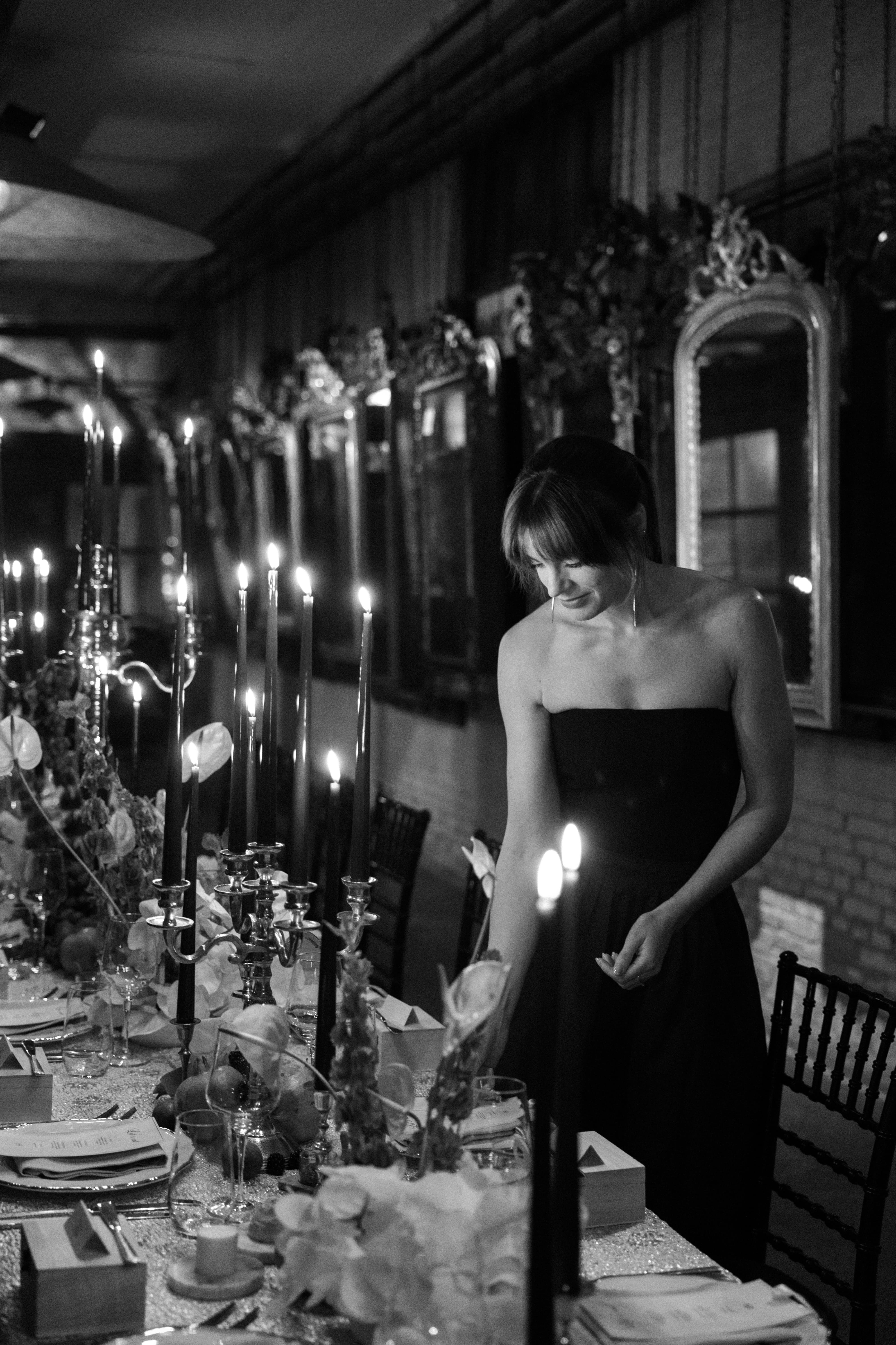 A woman in a strapless black dress stands beside a decorated dining table with many lit taper candles in a dimly lit indoor setting.