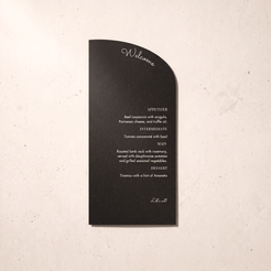 A black arched welcome placemat with white printed text, displayed flat against a textured off-white background.