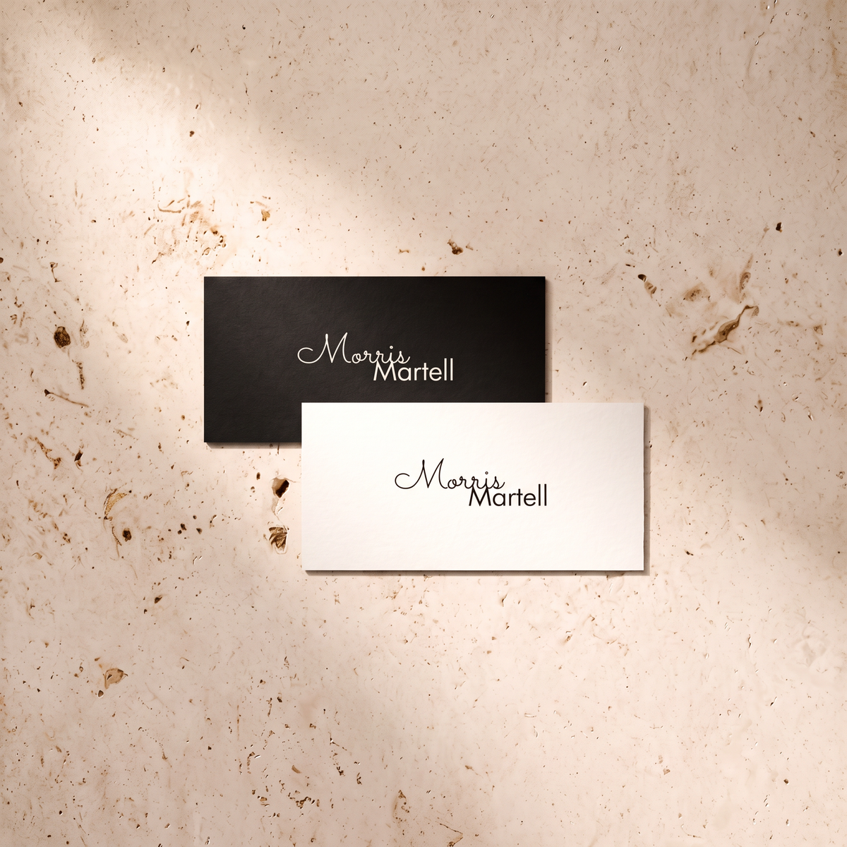 Two rectangular business cards with the name 'Morris Martell' printed on them, one black and one white, arranged on a textured beige stone surface with soft shadows.
