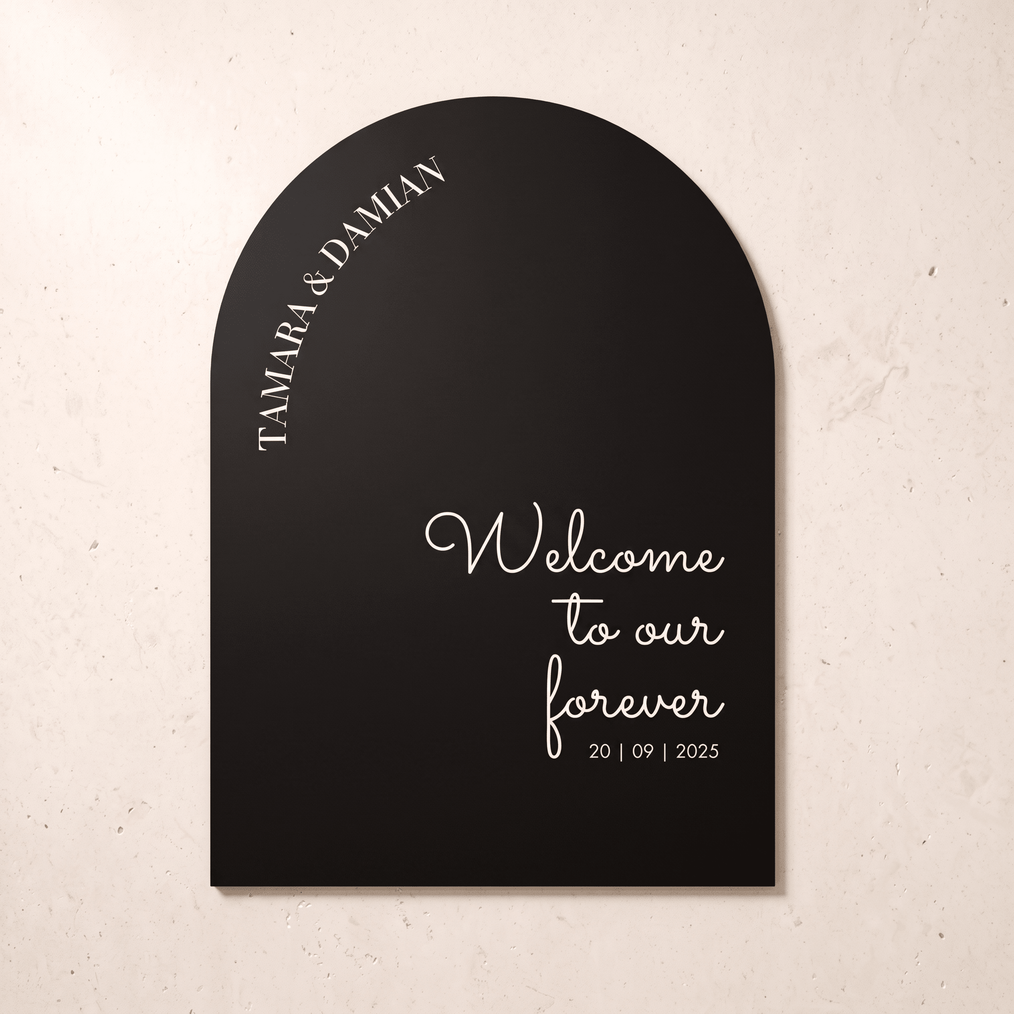 A black arched wall plaque with white script text reading 'Welcome to our forever 20 | 09 | 2025' and 'TAMARA & DAMIAN' curved along the left edge, shown against a light beige textured wall.