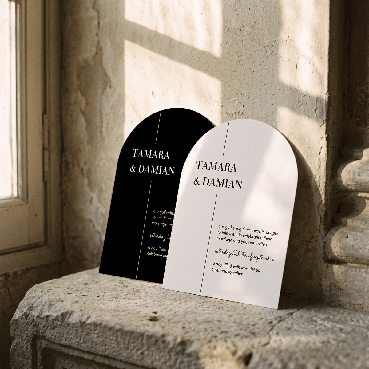 Two arched wedding invitation cards leaning against a stone wall in warm sunlight. One card is black with white text, and the other is white with black text.