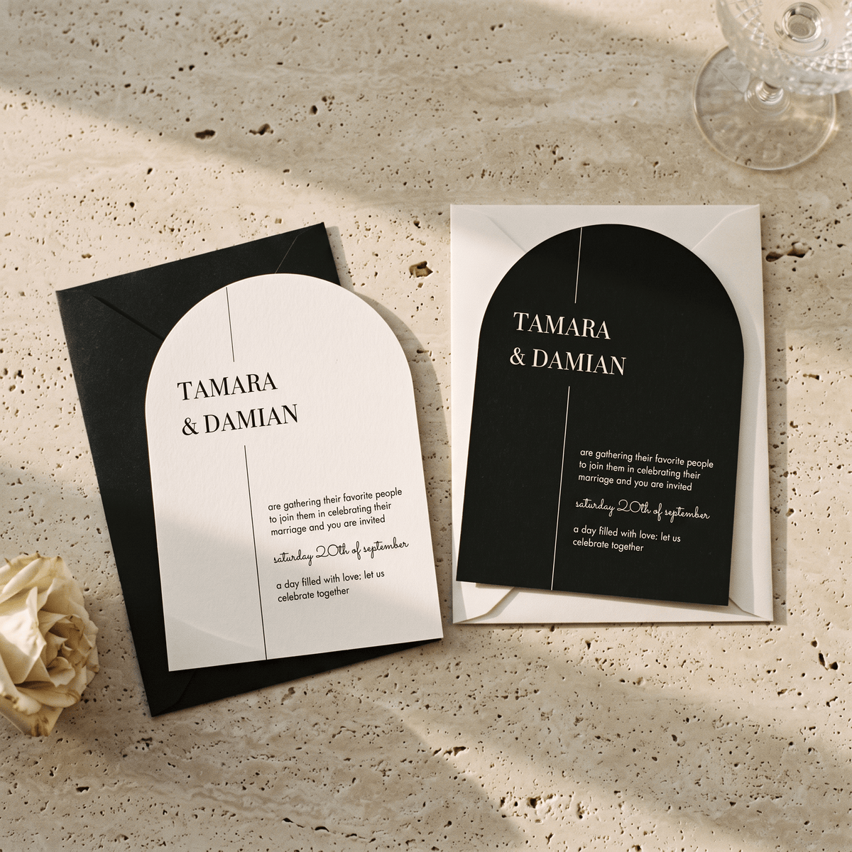 Architectural black and white wedding invitation suite with a matching black envelope, styled on a textured beige stone surface with a cream rose and glass stemware in the background.