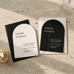 Architectural black and white wedding invitation suite with a matching black envelope, styled on a textured beige stone surface with a cream rose and glass stemware in the background.
