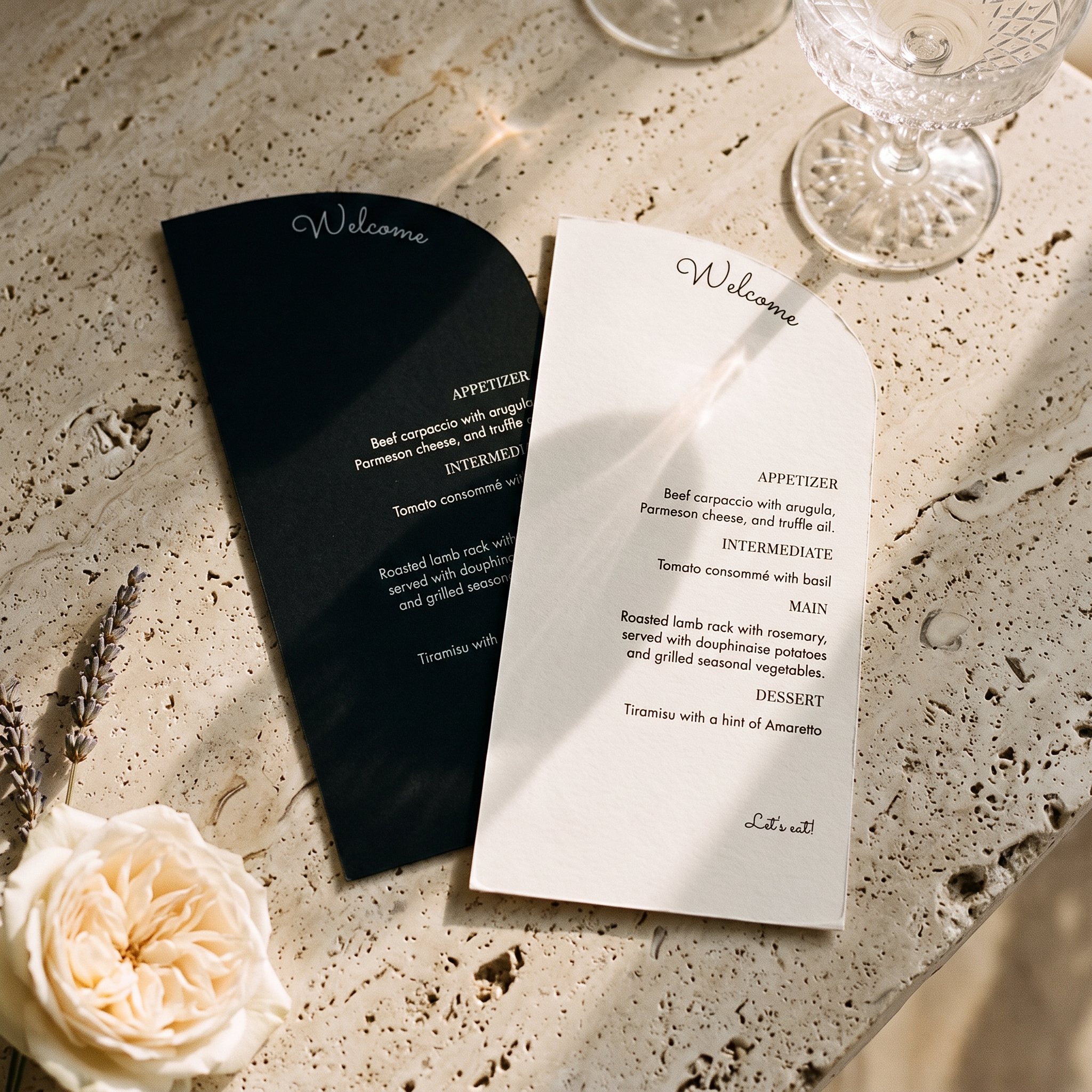 A wedding-style welcome card set styled on a textured stone surface, featuring a black card with white script and a white card with black text, surrounded by a cream rose, dried lavender, and a glass chalice.