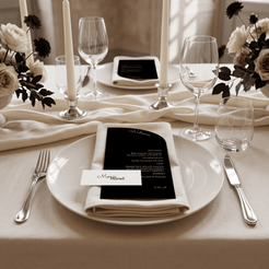A styled wedding table setting with a white plate, cream napkin, black place card, silver cutlery, clear wine glasses, and floral arrangements on a beige tablecloth.