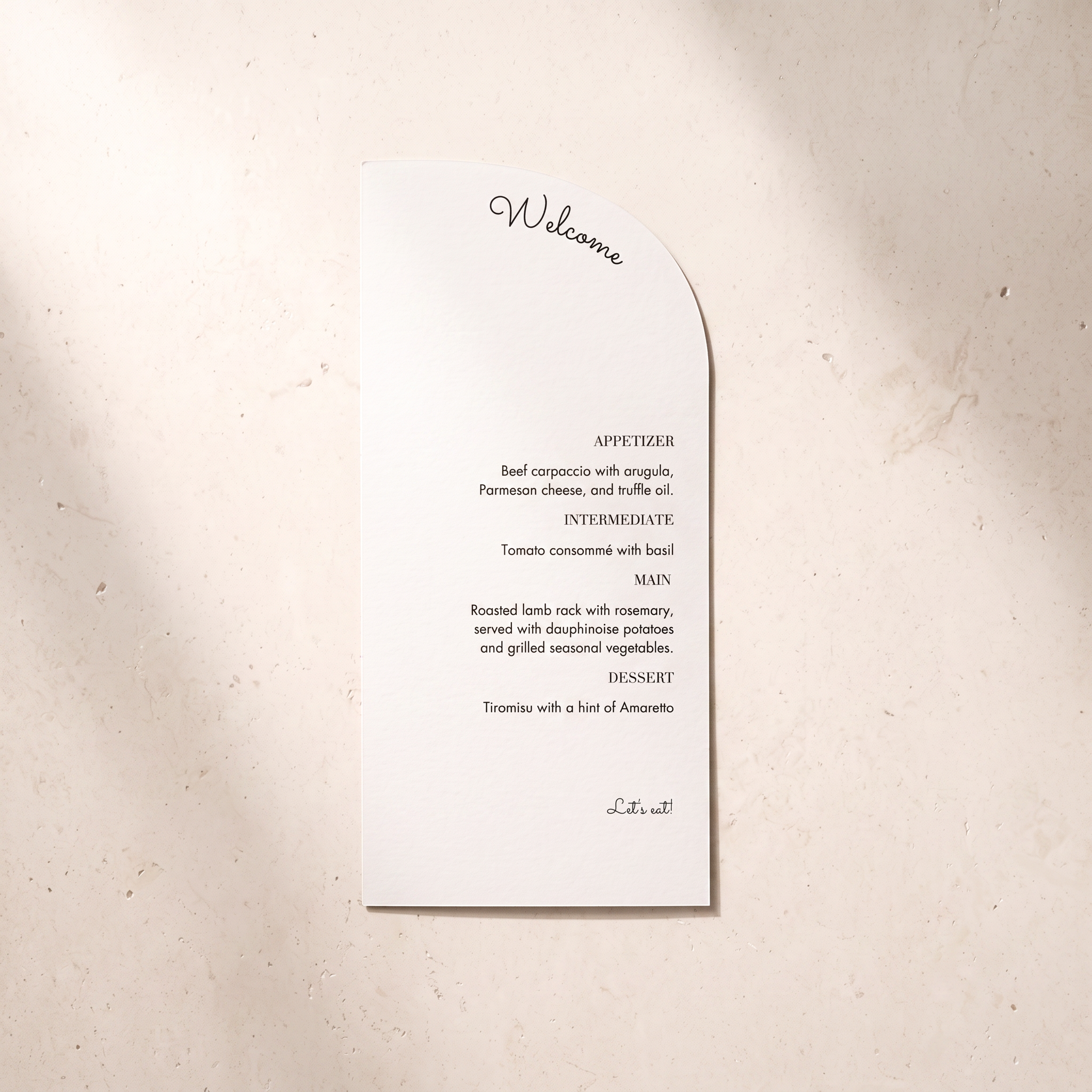 A white paper wedding program or invitation card with a curved right edge, printed with a welcome message and appetizer list in black text, placed on a light beige textured surface.