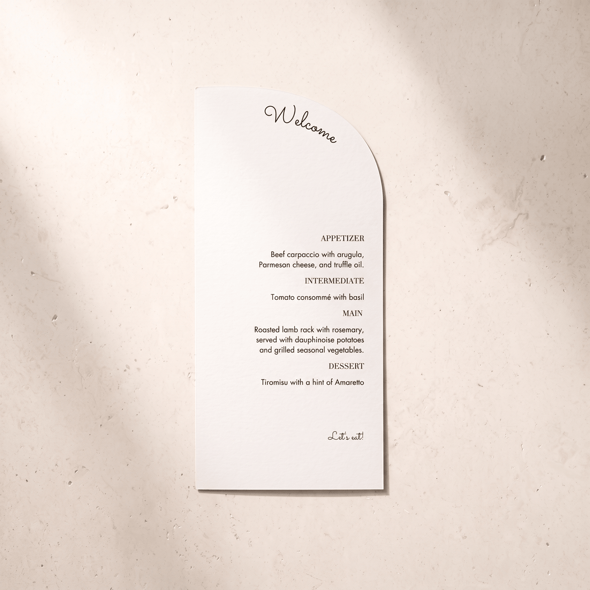 A white paper wedding program or invitation card with a curved right edge, printed with a welcome message and appetizer list in black text, placed on a light beige textured surface.