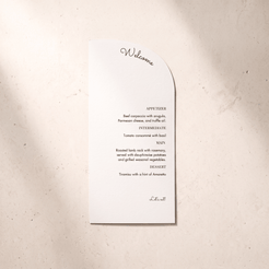 A white paper wedding program or invitation card with a curved right edge, printed with a welcome message and appetizer list in black text, placed on a light beige textured surface.