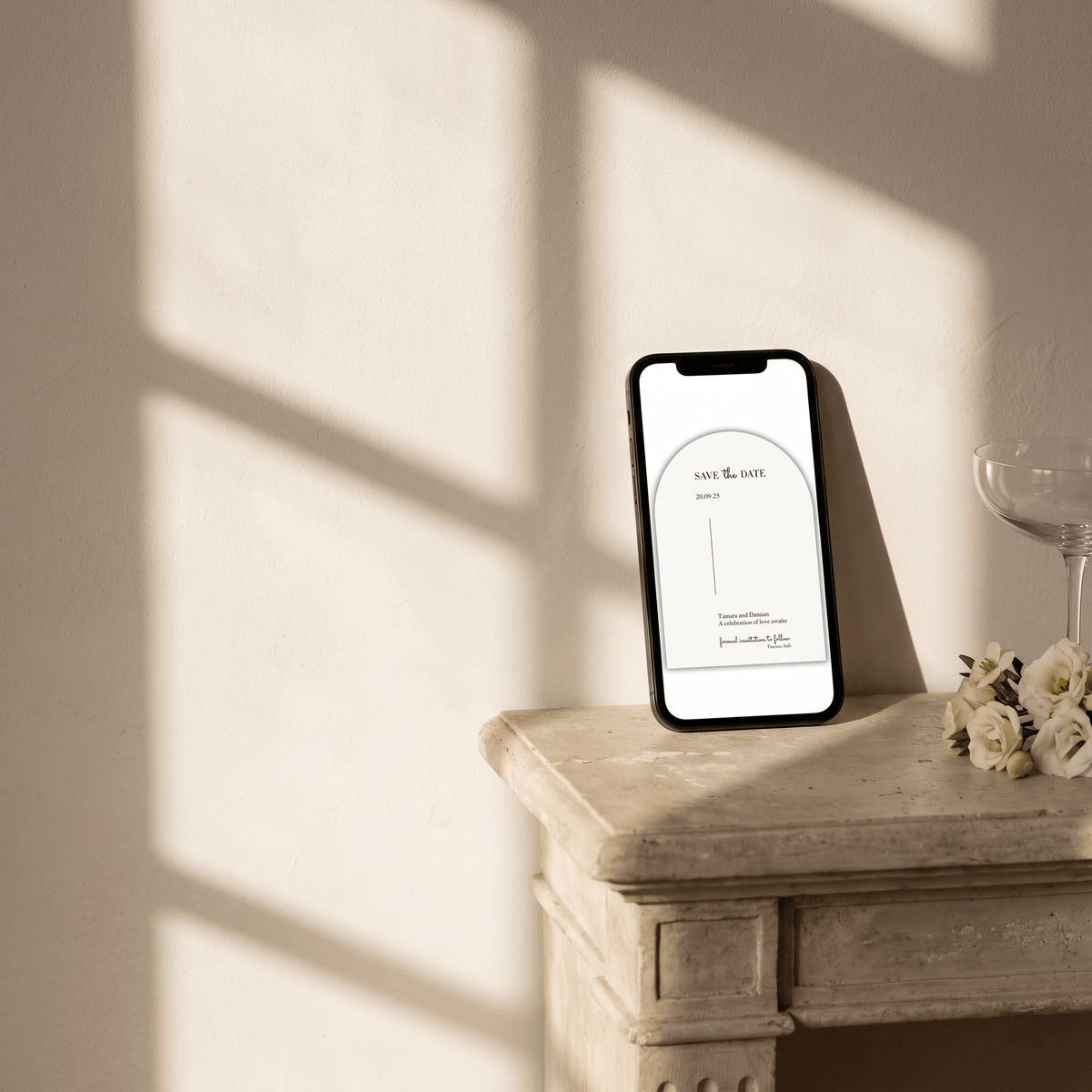 A smartphone displaying a minimalist wedding invitation mockup stands on a rustic stone table beside a wine glass and small white flowers, with warm sunlight casting window-frame shadows on a cream wall.