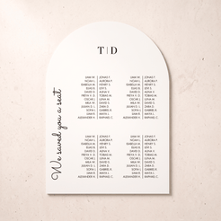 An arched white wedding seating card with black printed text, featuring the heading 'T | D' at the top, a list of names under that heading, and the phrase 'We saved you a seat' in script on the left side, photographed on a light beige textured background.