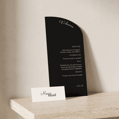 A black arched welcome sign with white text stands on a light stone shelf next to a small white name card reading 'Maggie Marrell' against a warm beige wall.