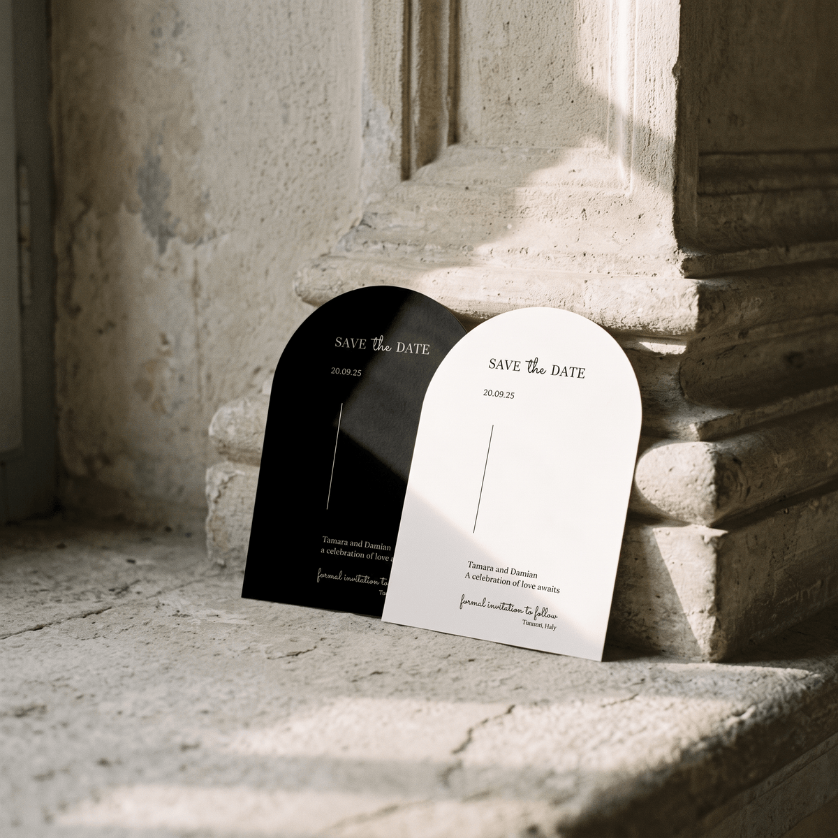 Two arched 'Save the Date' cards leaning against a stone ledge in natural sunlight. One card is black with white text, and the other is white with black text, set against a rustic stone wall and column.