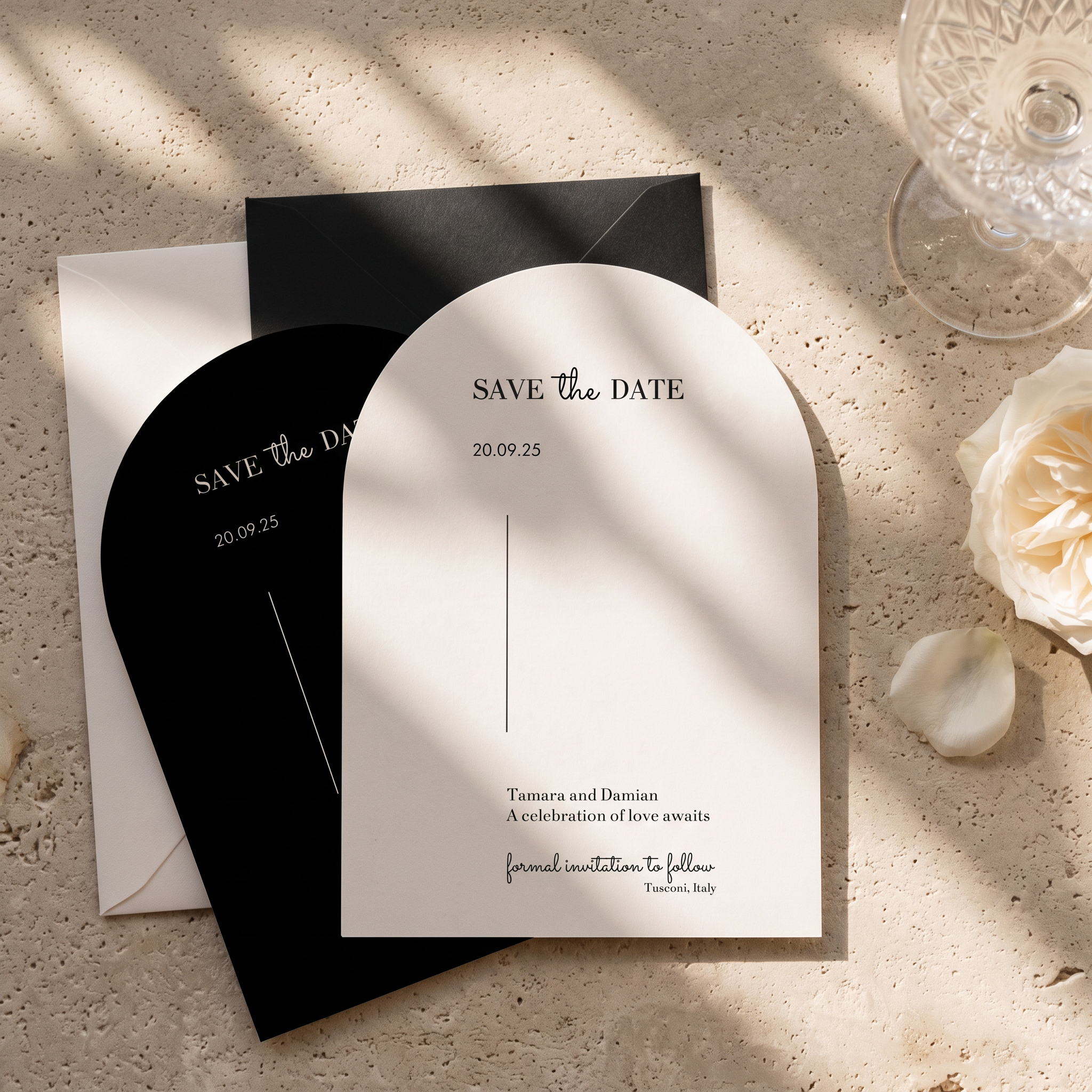 Arch-shaped white and black 'Save the Date' wedding stationery cards arranged on a textured beige stone surface with soft window shadows, alongside a glass and a white rose.