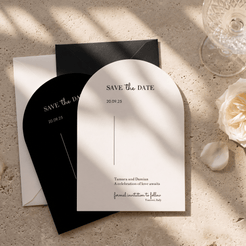Arch-shaped white and black 'Save the Date' wedding stationery cards arranged on a textured beige stone surface with soft window shadows, alongside a glass and a white rose.