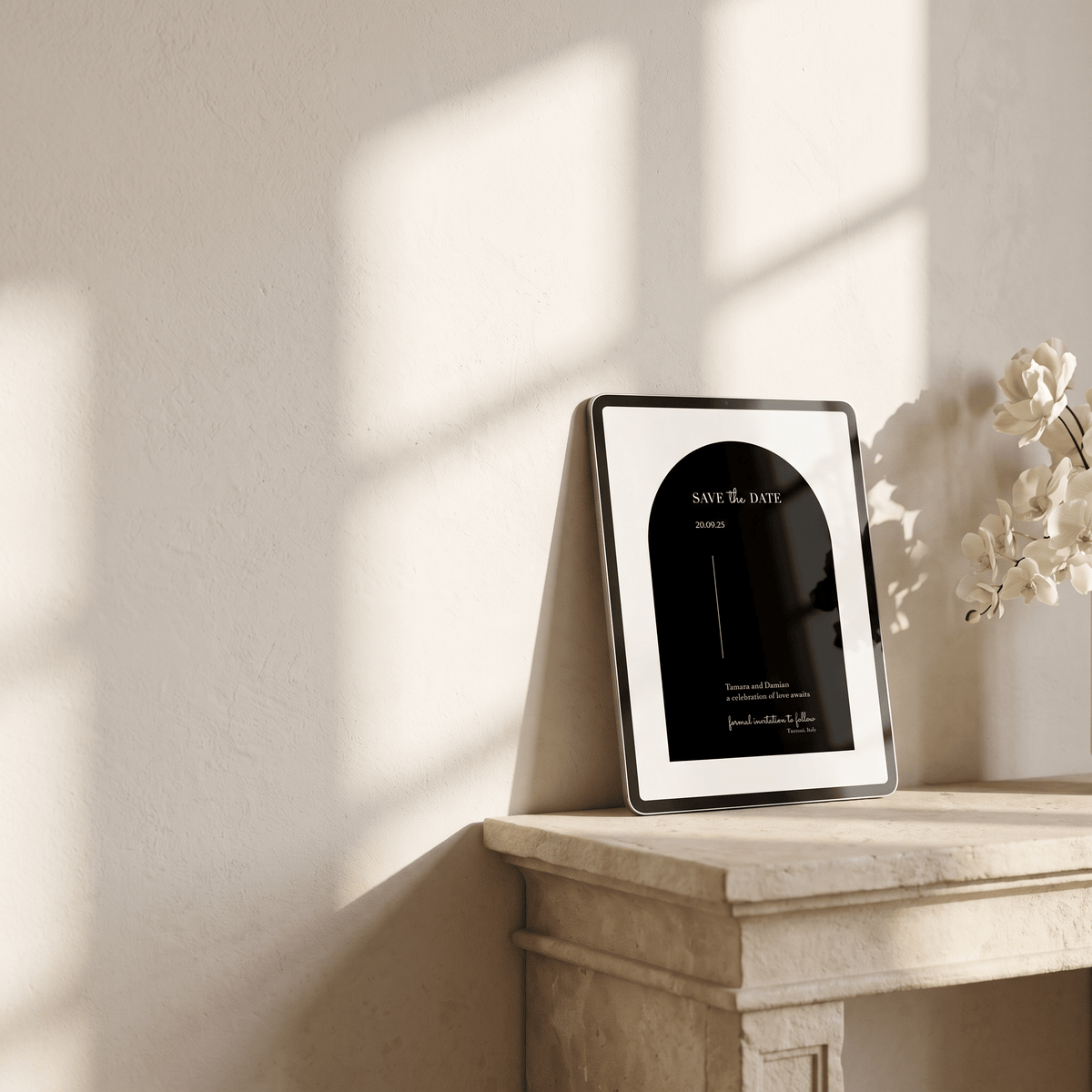 A framed minimalist black and white art print with 'SAVE THE DATE' text, leaning against a textured beige wall on a stone mantelpiece with soft window shadows and white flowers nearby.