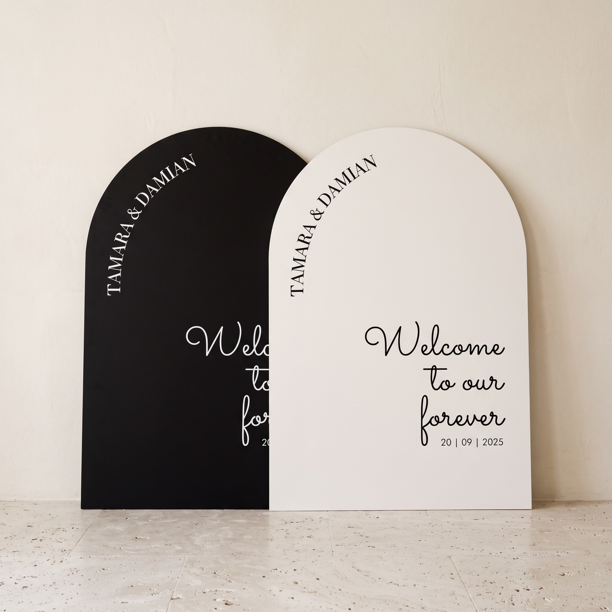 Two arched welcome sign panels leaning against a light beige wall on a stone surface. One panel is matte black with white script text reading 'Welcome to our forever' and the other is matte white with black script text reading 'Welcome to our forever.