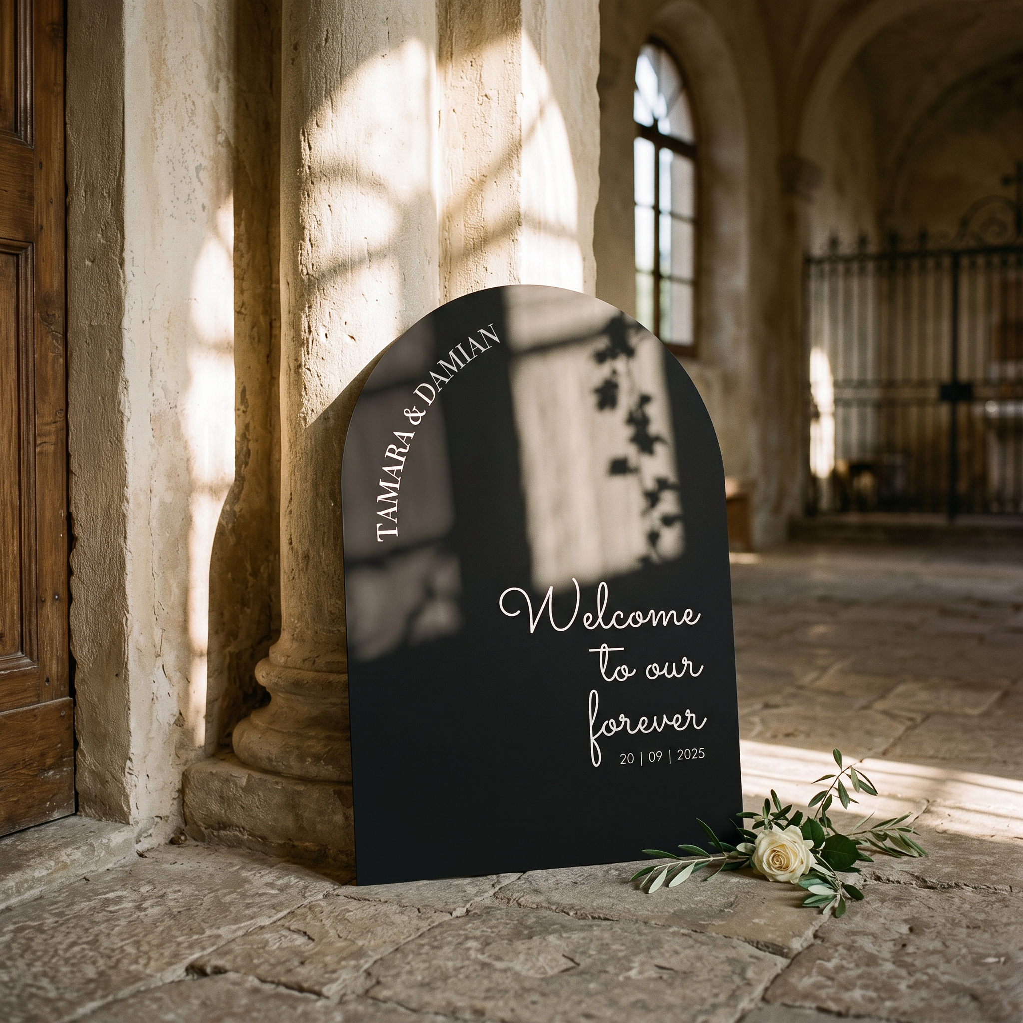 A black arched wedding welcome sign with white script text reading 'Welcome to our forever 20 | 09 | 2025' and 'TAMARA & DAMIAN' along the top edge, leaning against a stone column in a sunlit historic interior with a small white rose arrangement at the base.